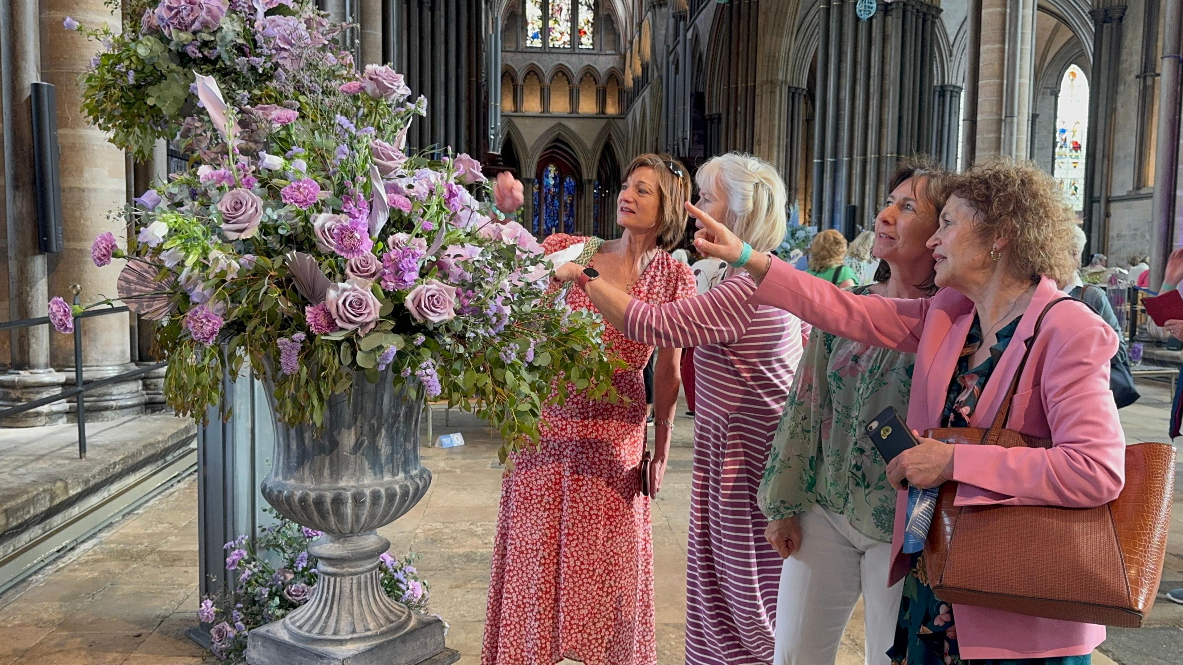 A arrangement of pink and purple flowers in a stone vase in the middle of the cathedral's main hall. Four women wearing pink and red clothes are looking and pointing at the flowers