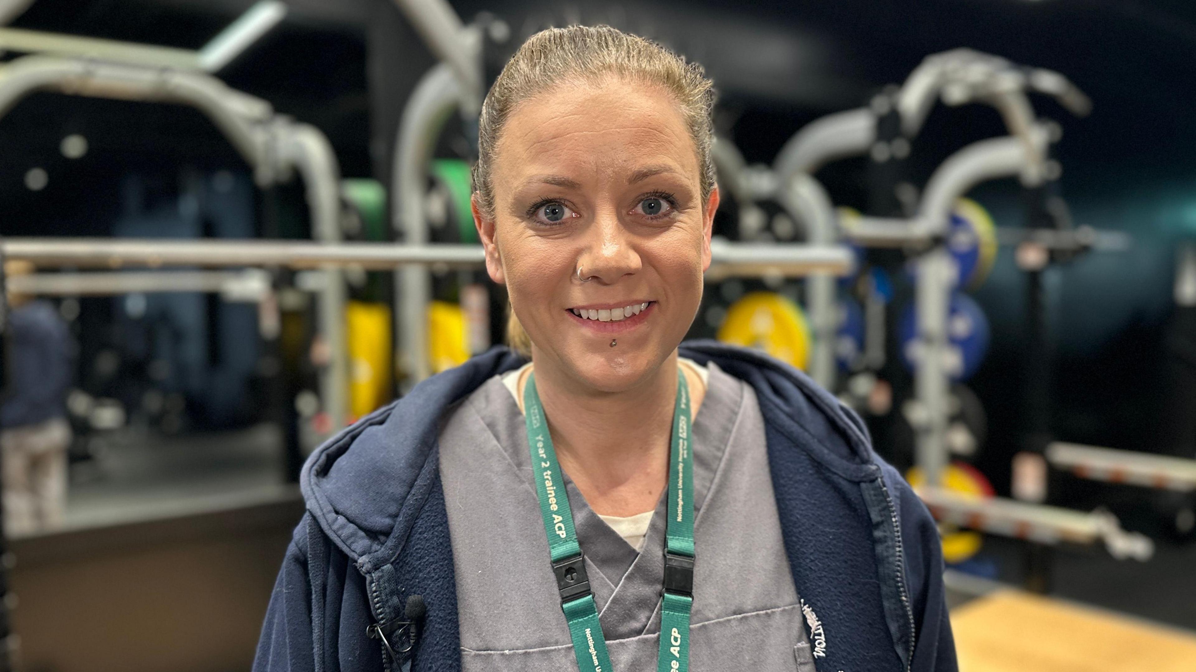 Naomi Richmond, dressed in her hospital uniform, smiling at the camera while stood in front of gym equipment