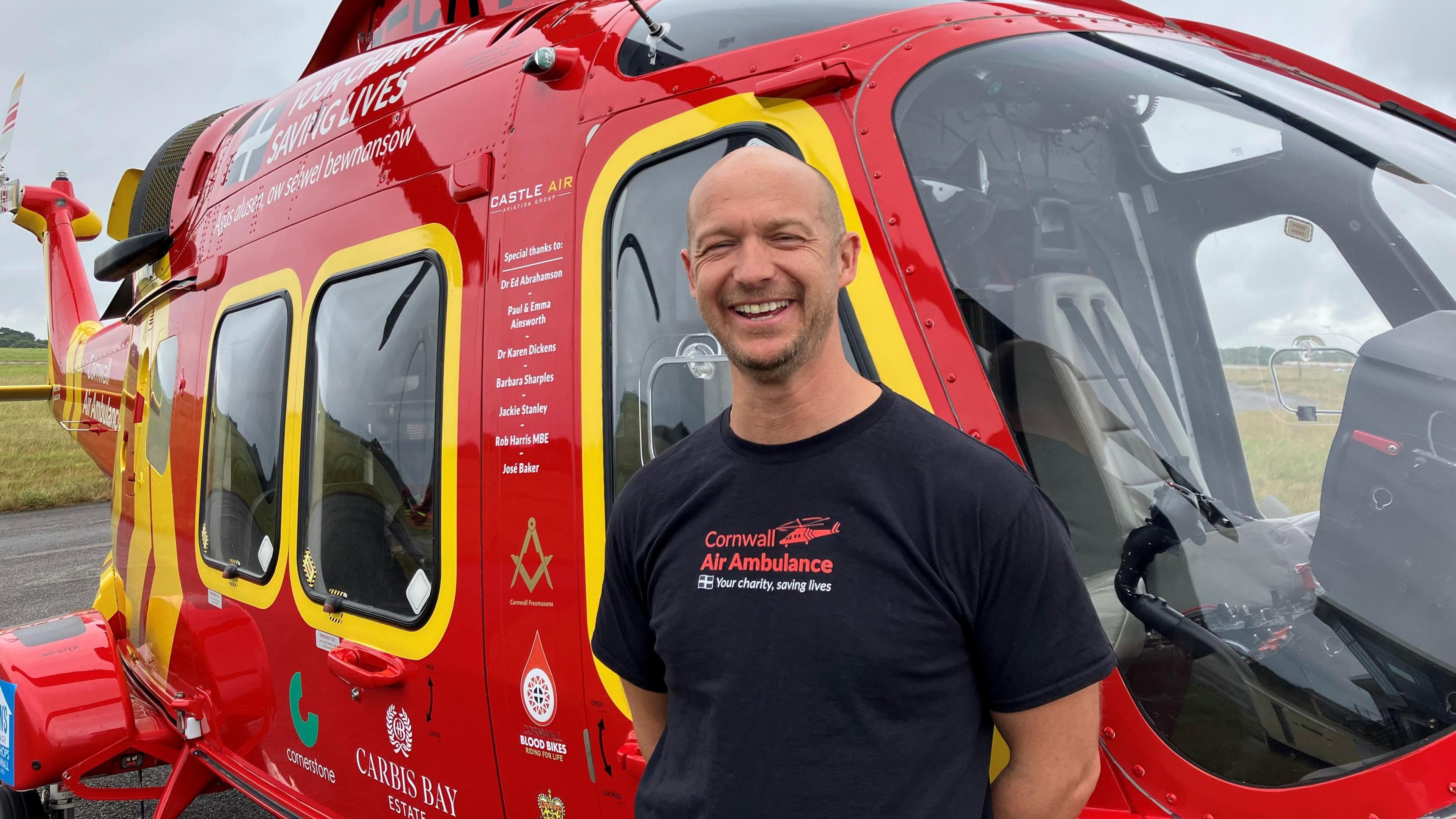 Captain Adam Smith is laughing as he stands in front of the air ambulance. He has his hands held behind his back and is wearing a branded Cornwall Air Ambulance T-shirt. He has stubble and a shaved head.