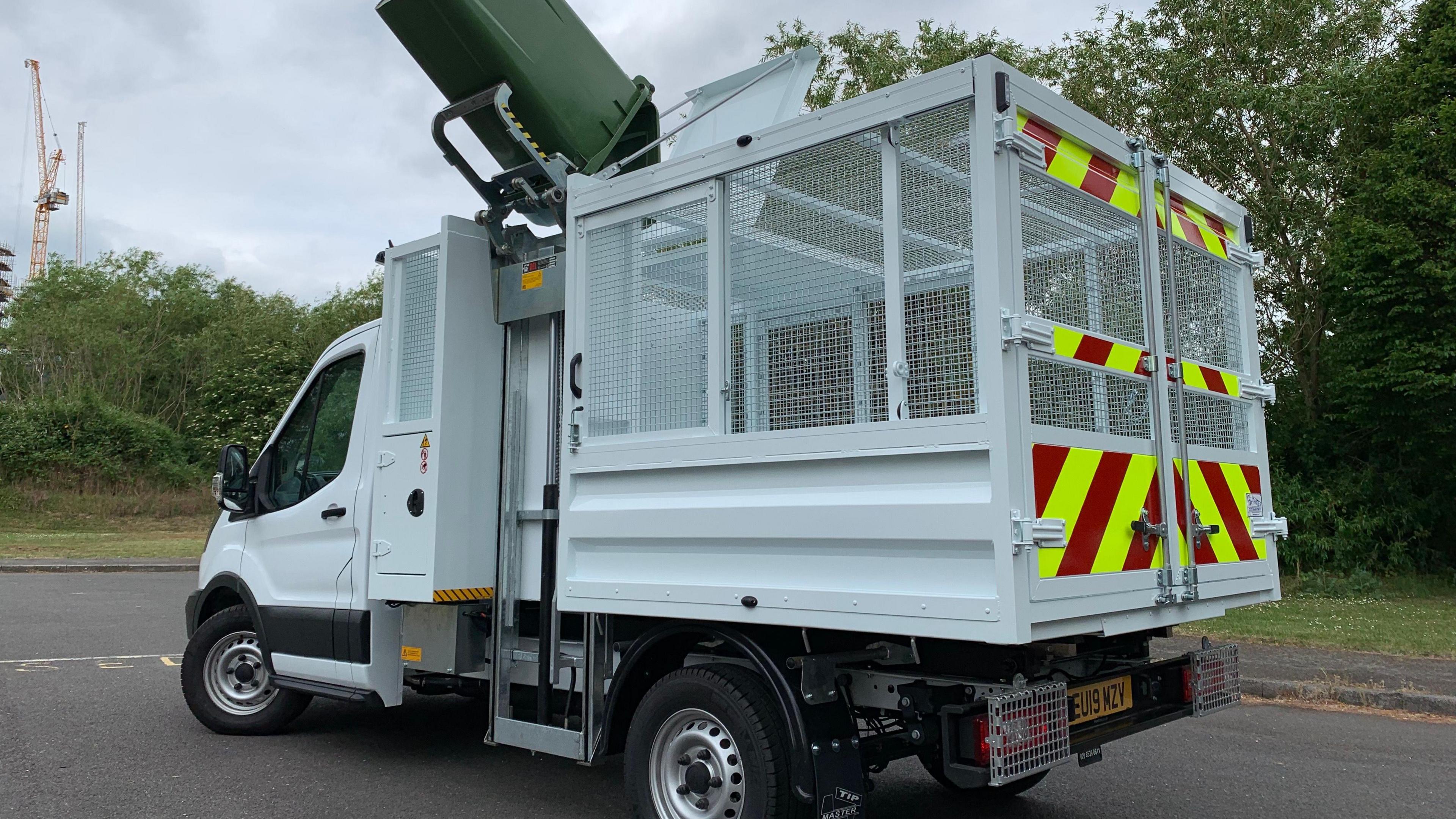 A small lorry with an enclosed cage on the back, which has a bin carrying lift on the side which accesses a hatch on top.