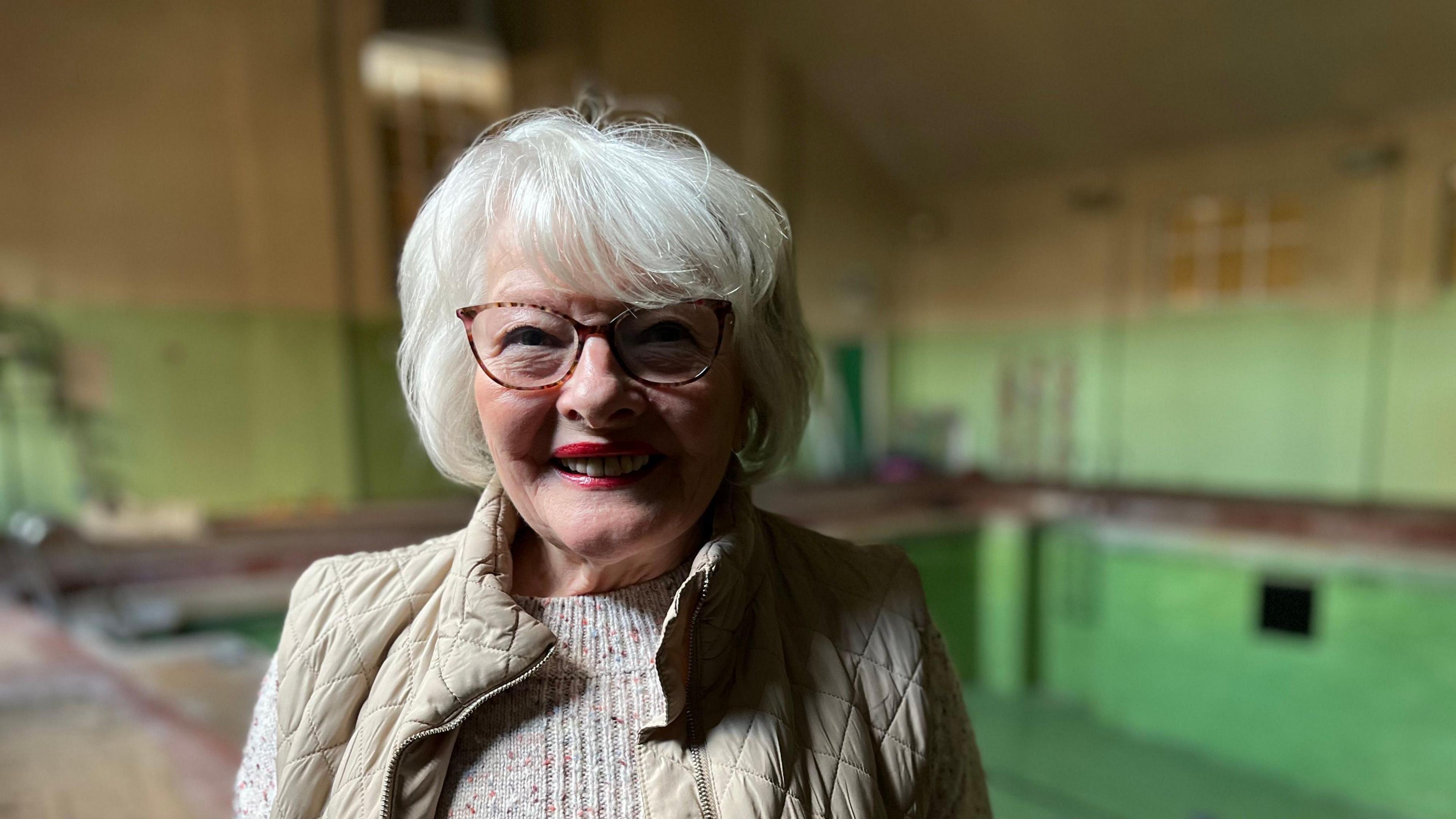 A woman wearing glasses, a beige jumper and a cream gilet stands in front of an empty swimming pool.