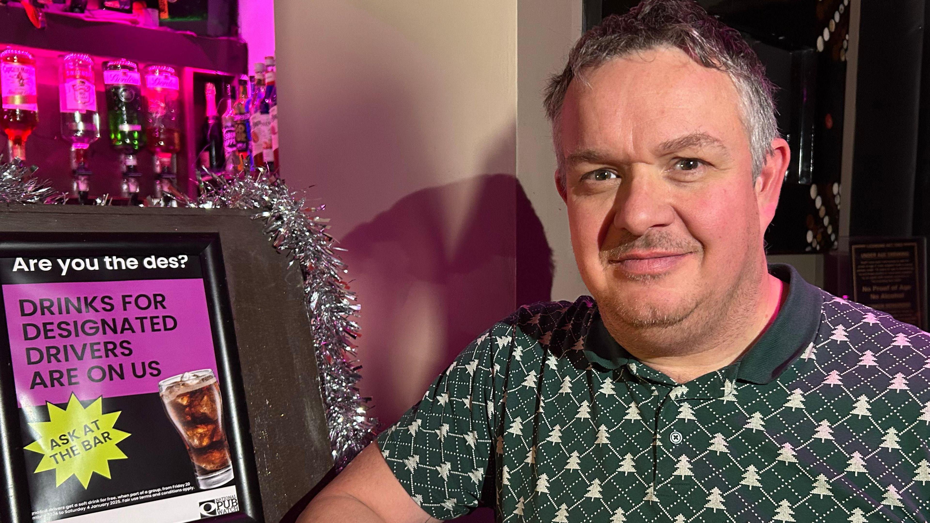 A man in a green polo shirt with Christmas trees on stands next to a bar with a poster displaying the offer of free drinks for designated drivers. 