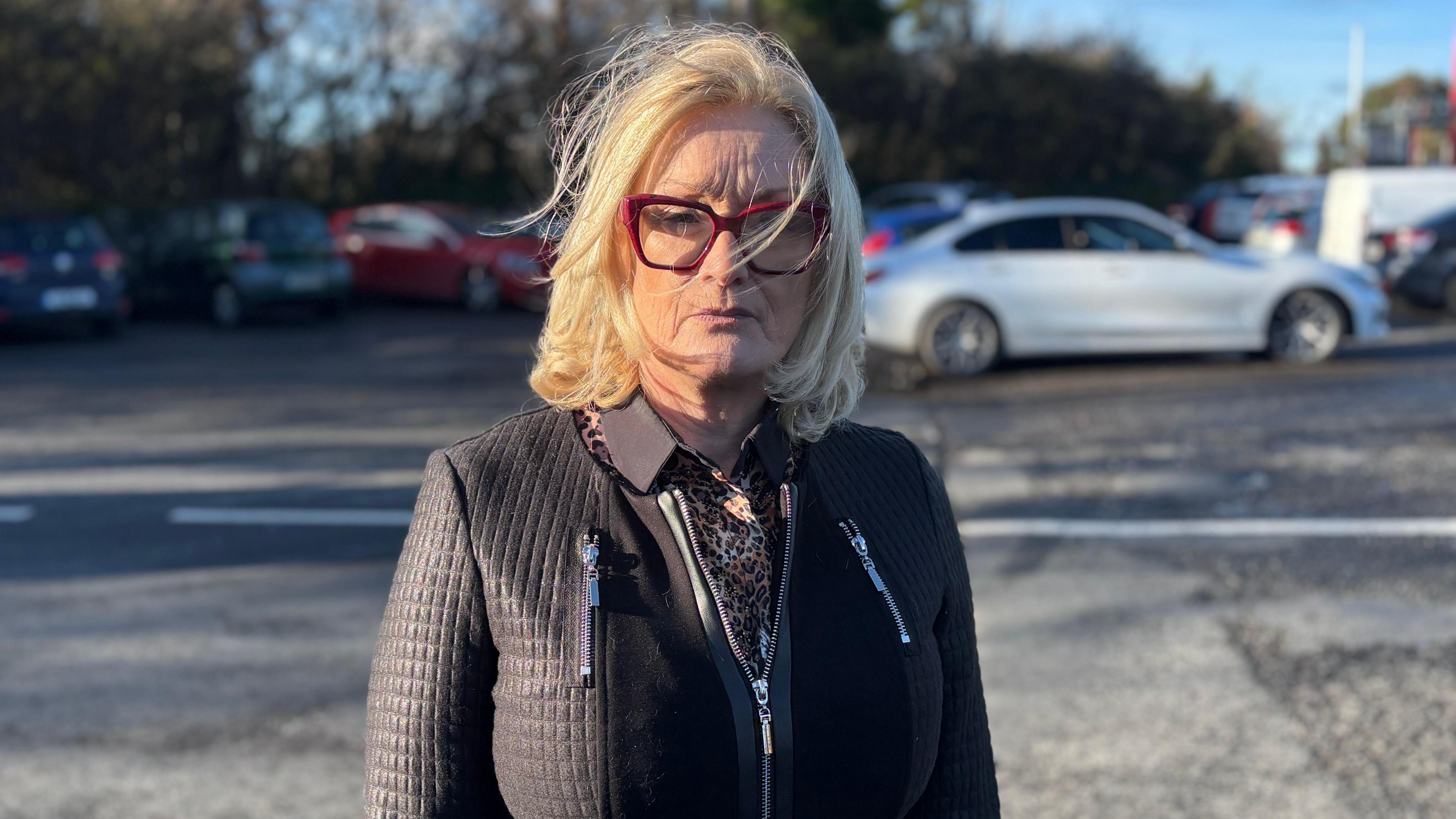 Sharon is looking at the camera with a serious expression. She has red glasses and shoulder length blonde hair. She is wearing a cheetah print shirt under a black zip up jacket.