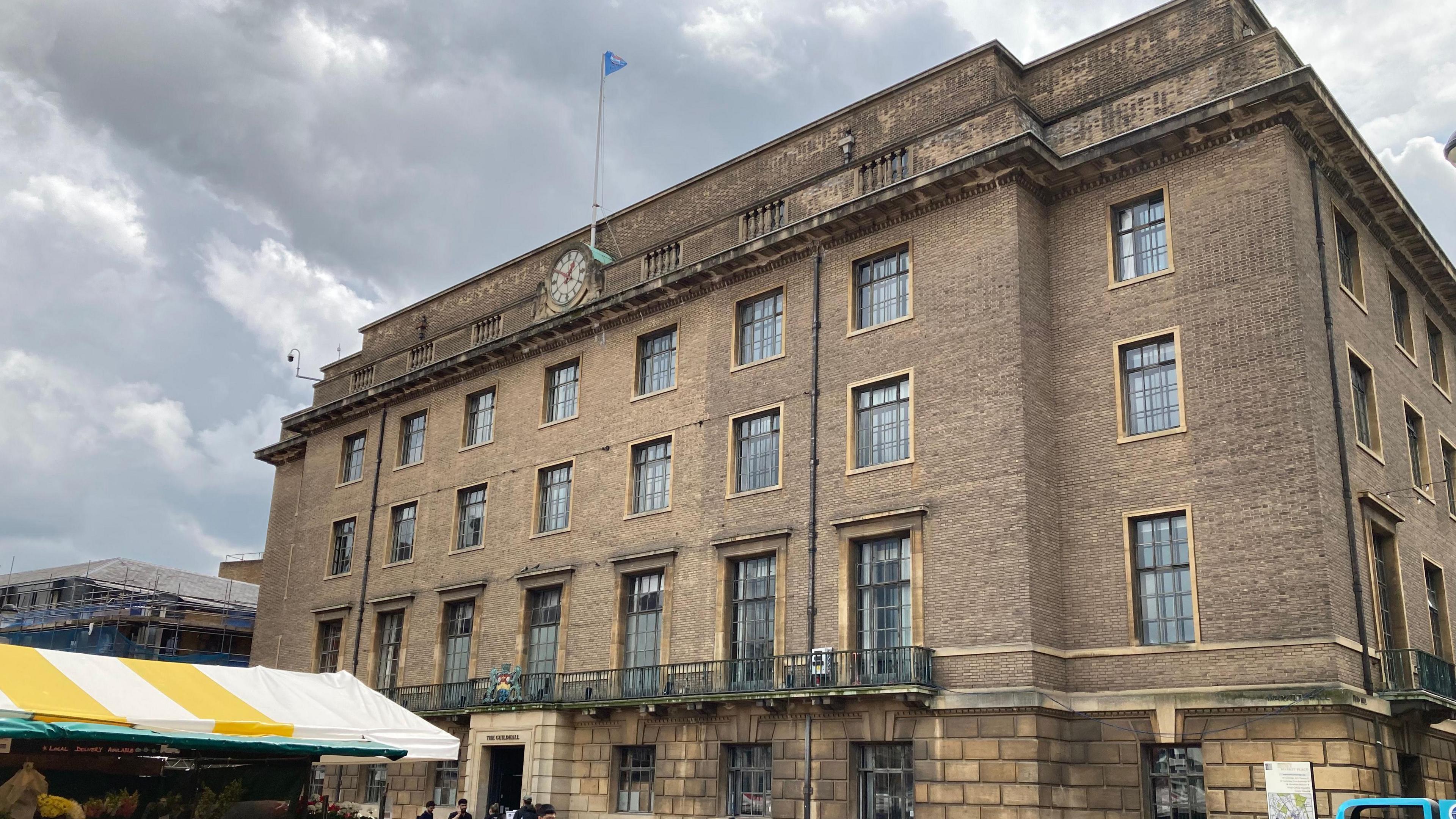 A 4 story building with a balcony on the 1st floor, a clock at the top and a flag flying above. A market stall is visible on the left hand side.
