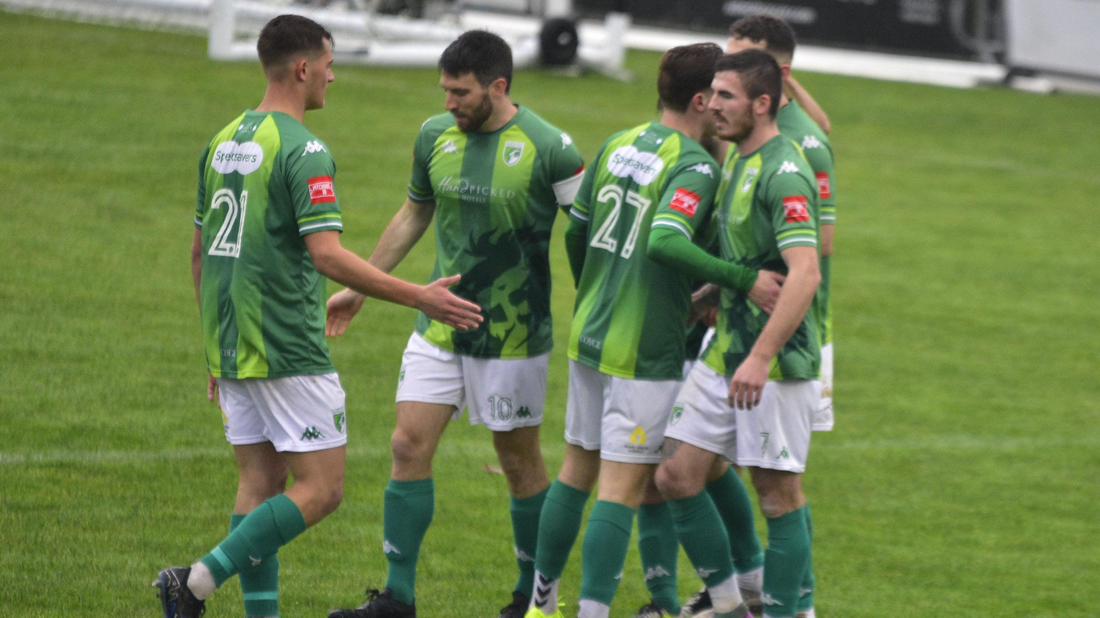 Six Guernsey FC players celebrate with each other after scoring a goal. All are wearing the football club's green home kit with white shorts and green socks.