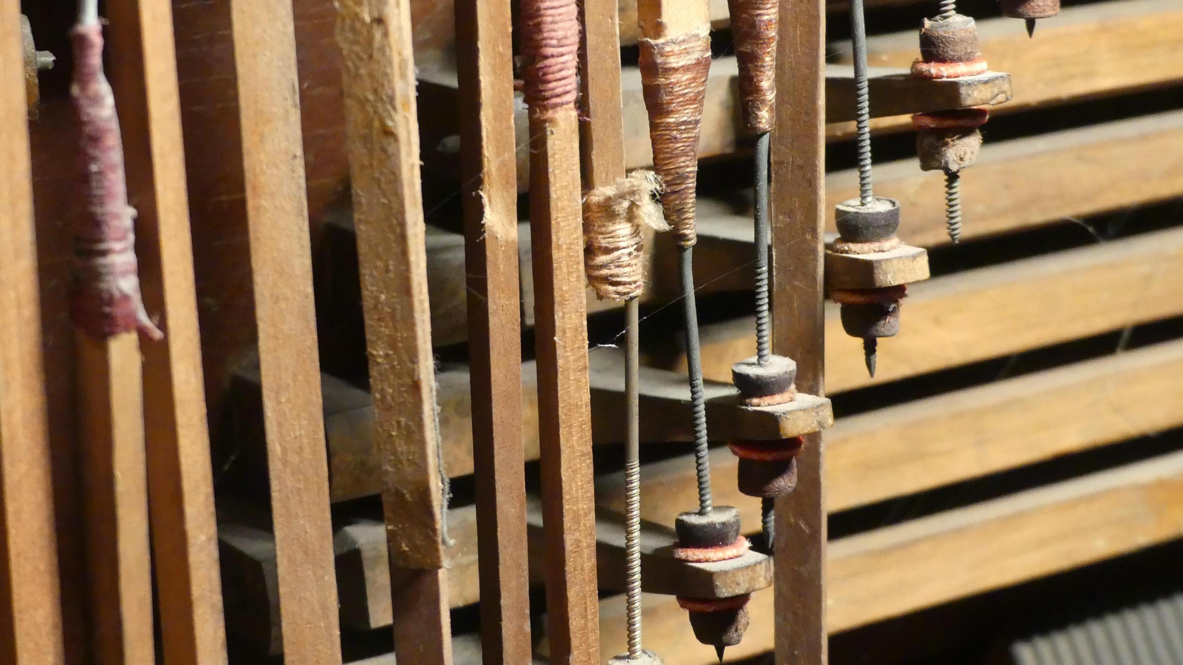 Worn internal workings inside an organ, comprised of wooden stakes wrapped in twine with metal hooks. Some of the hooks are loose and rough repairs have been carried out on the twine.