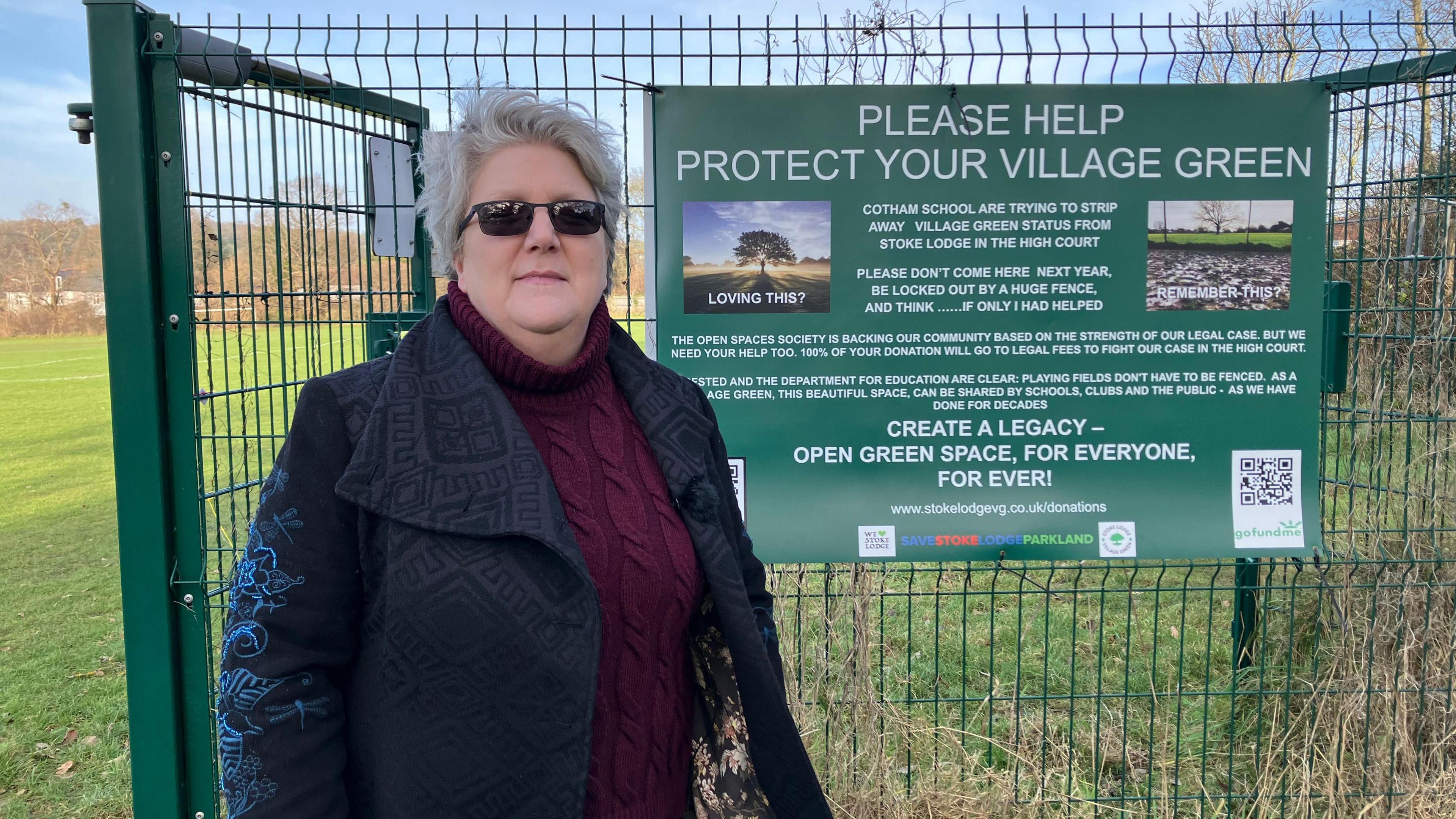 A woman wearing a black jacket, burgundy jumper and black sunglasses stands in front of a tall green fence with a sign saying: 'Please help protect your village green.'