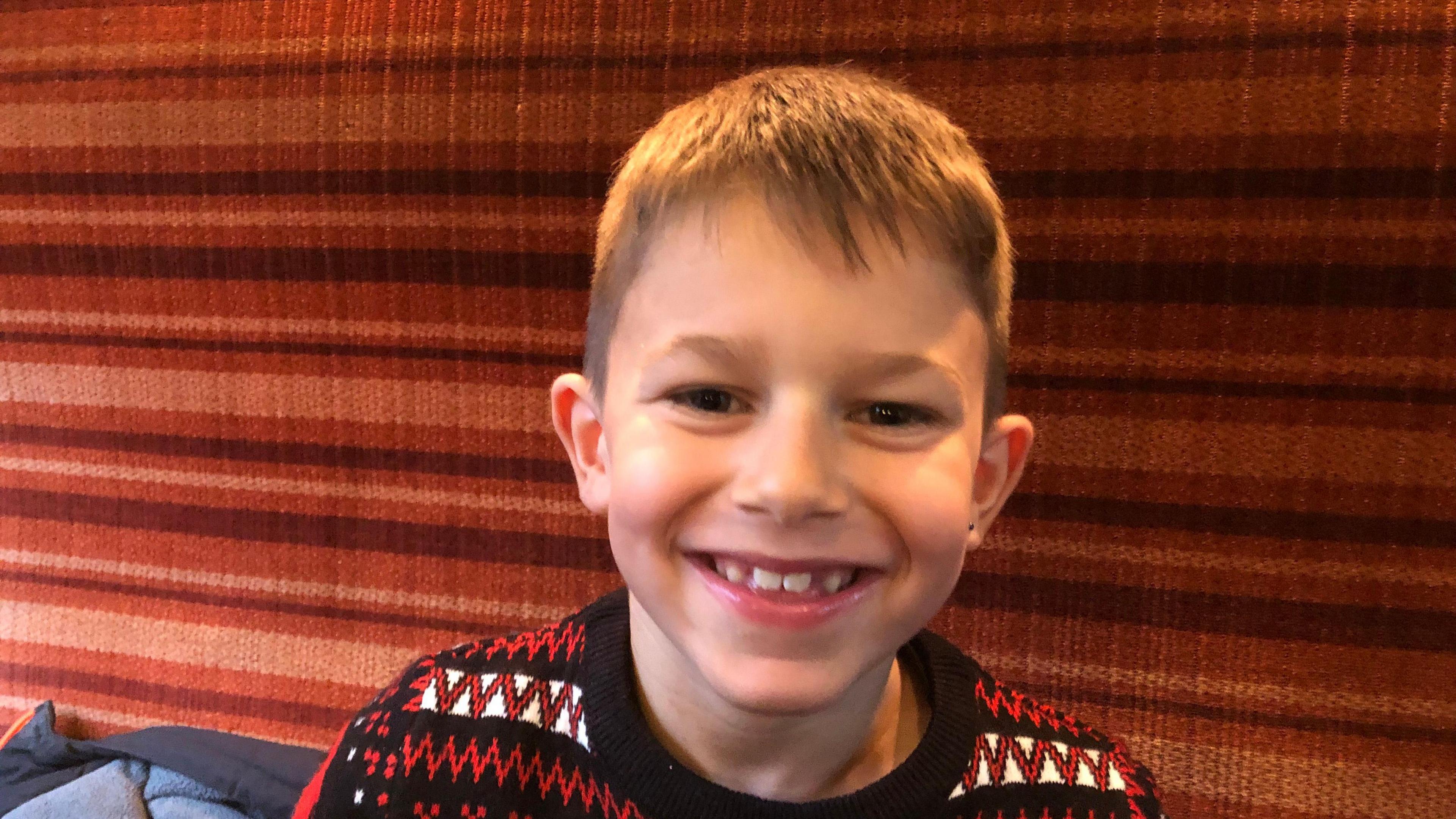 Zayden has short, light brown hair and is wearing a red, black and white Christmas jumper. He is smiling as his sits in a railway compartment