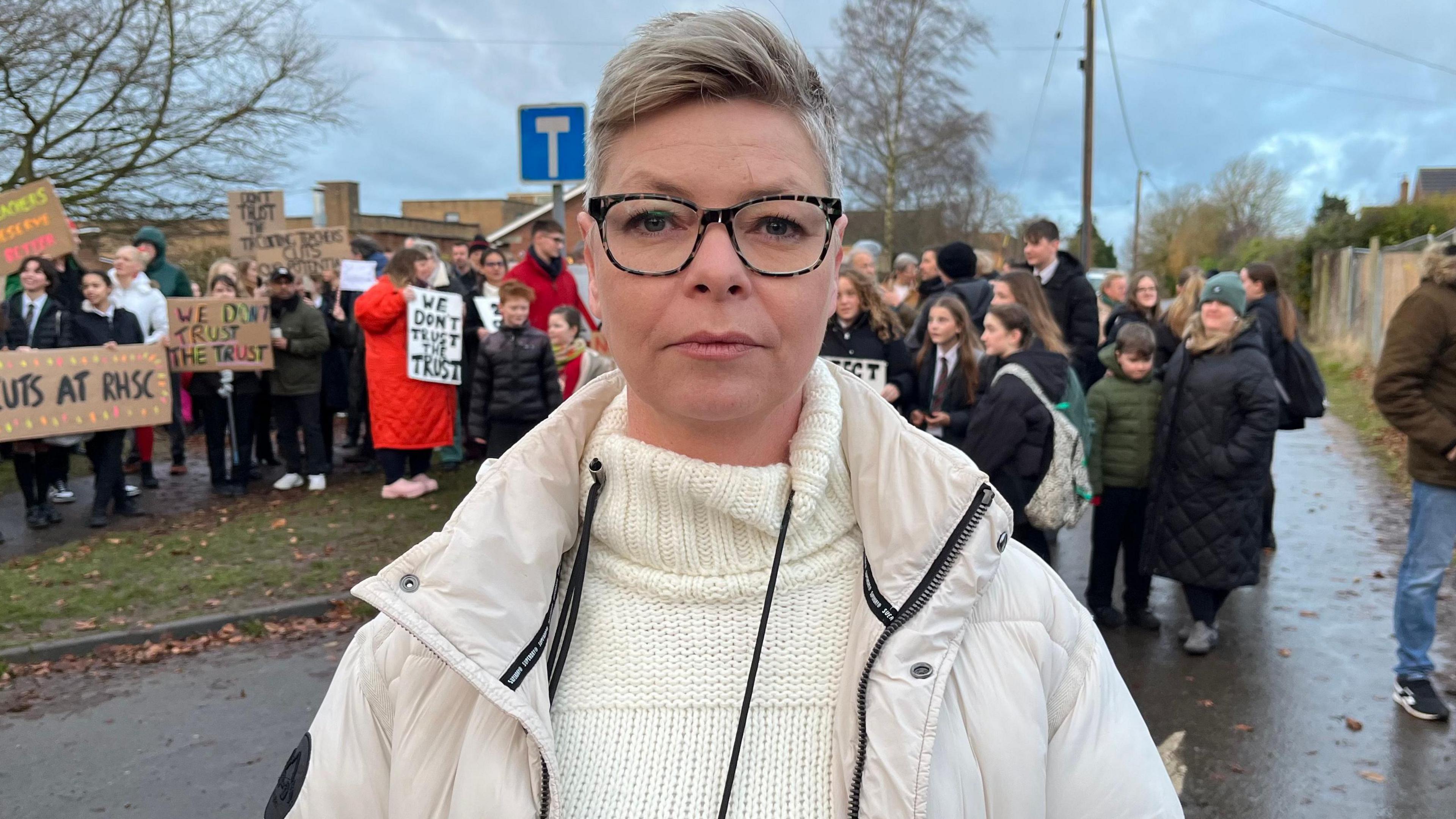 Laura Marfell-Williams, with swept back short hair is wearing glasses and a white puffer-style coat and cream jumper. Behind her are a group of protesters, some holding placards.