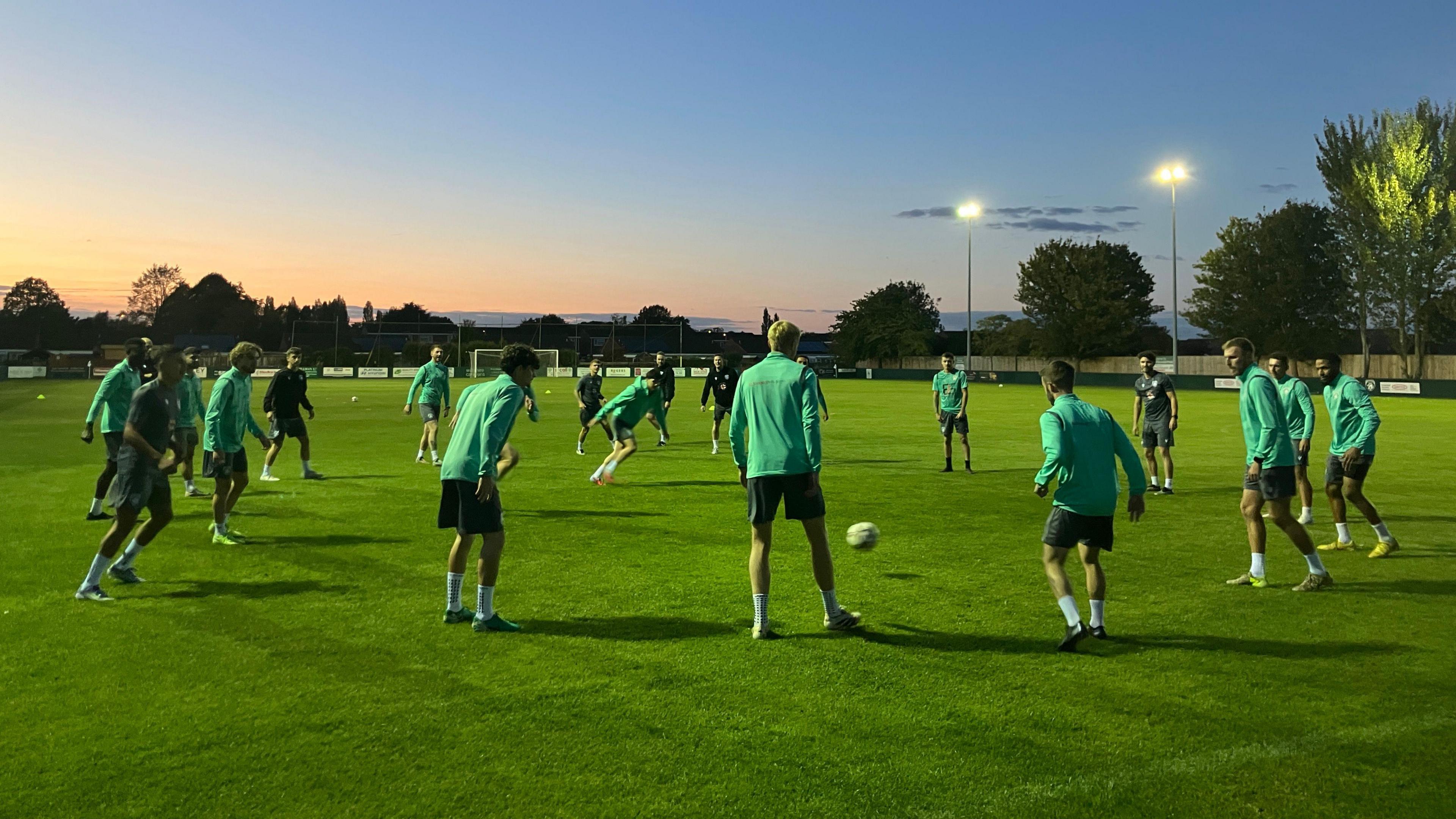 Westbury United players training in a circle as the sun goes down. The players are all wearing green tracksuits, apart from two who are wearing dark t-shirts and dark shorts. Two lit floodlights and the tree line can be seen in the background.