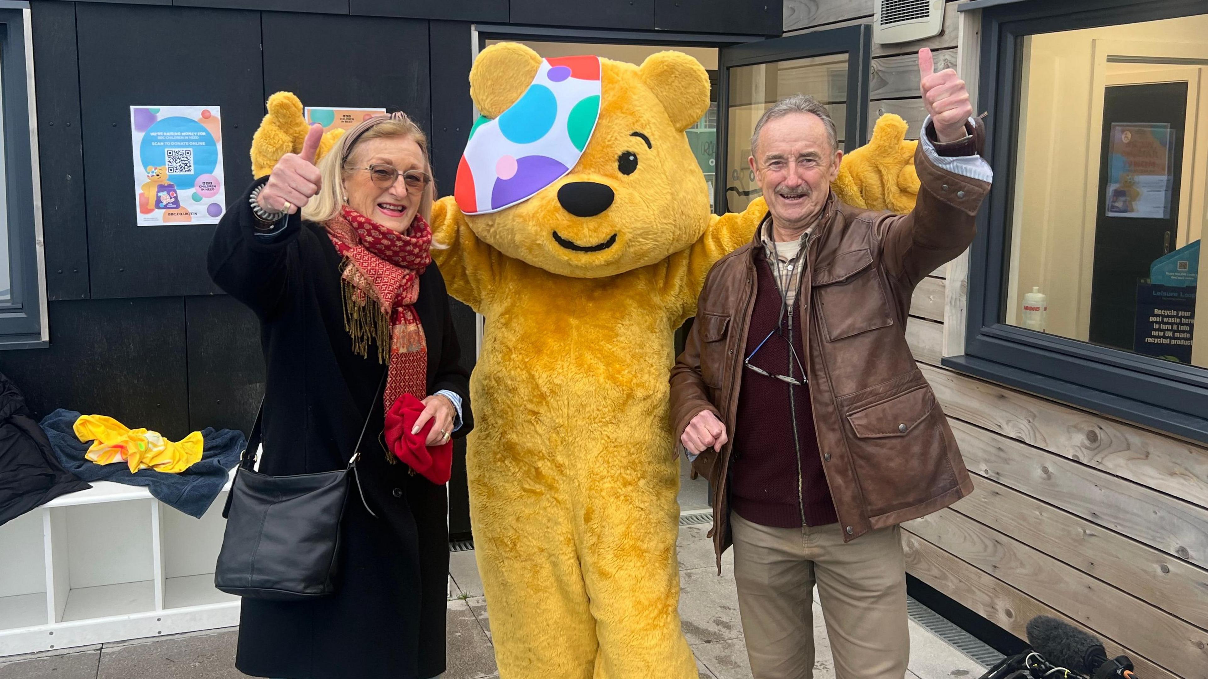 A woman with a long black coat on and a red scarf and a man with a brown leather jacket on smiling and looking at the camera. They are both holding up one 'thumbs up' in the air. They are either side of a person dressed as Pudsey.