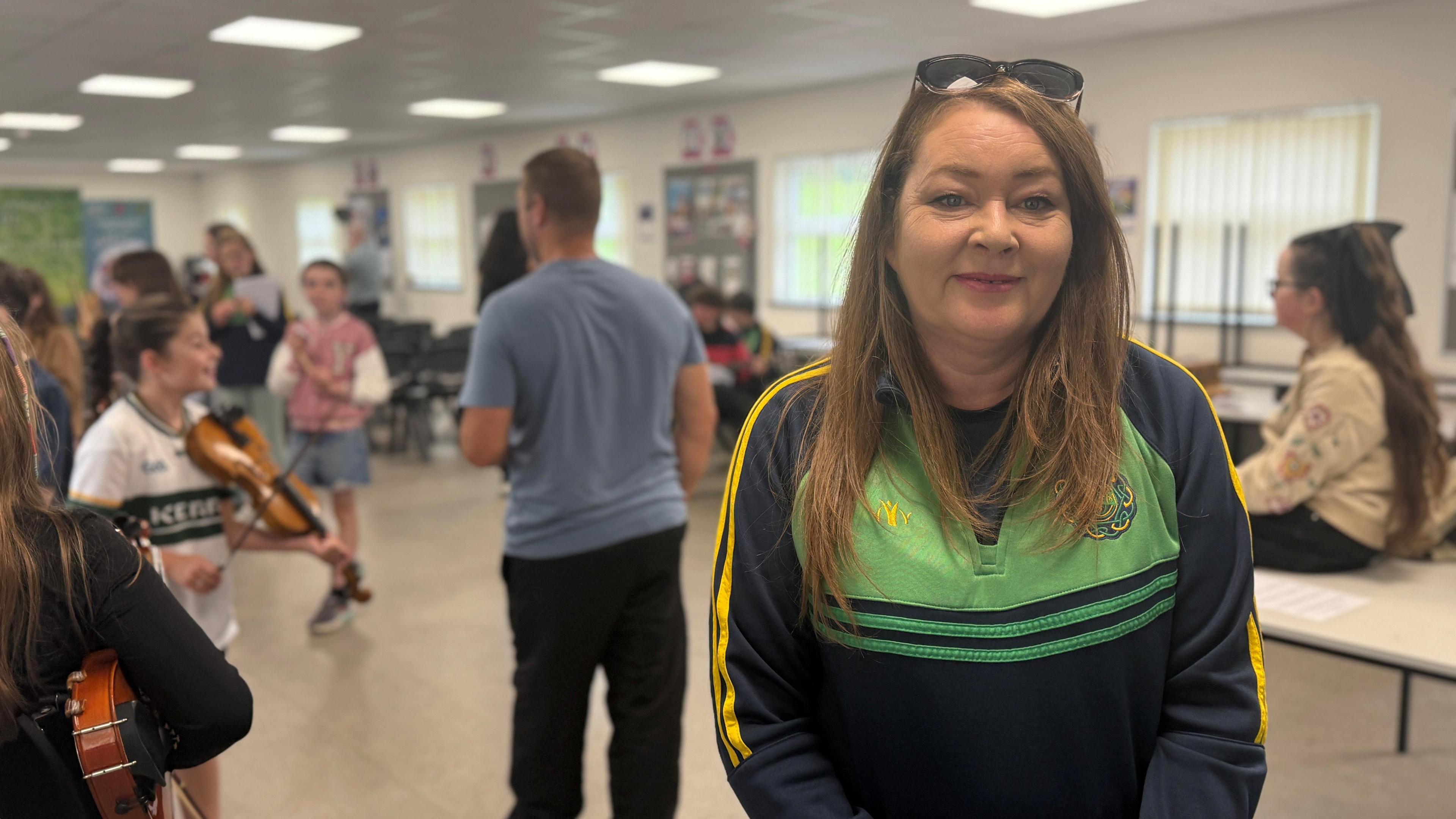 A woman with brown hair, with glasses perched on her head, wearing a navy and green uniform top. She is pictured in a busy classroom with multiple children in the background holding instruments.