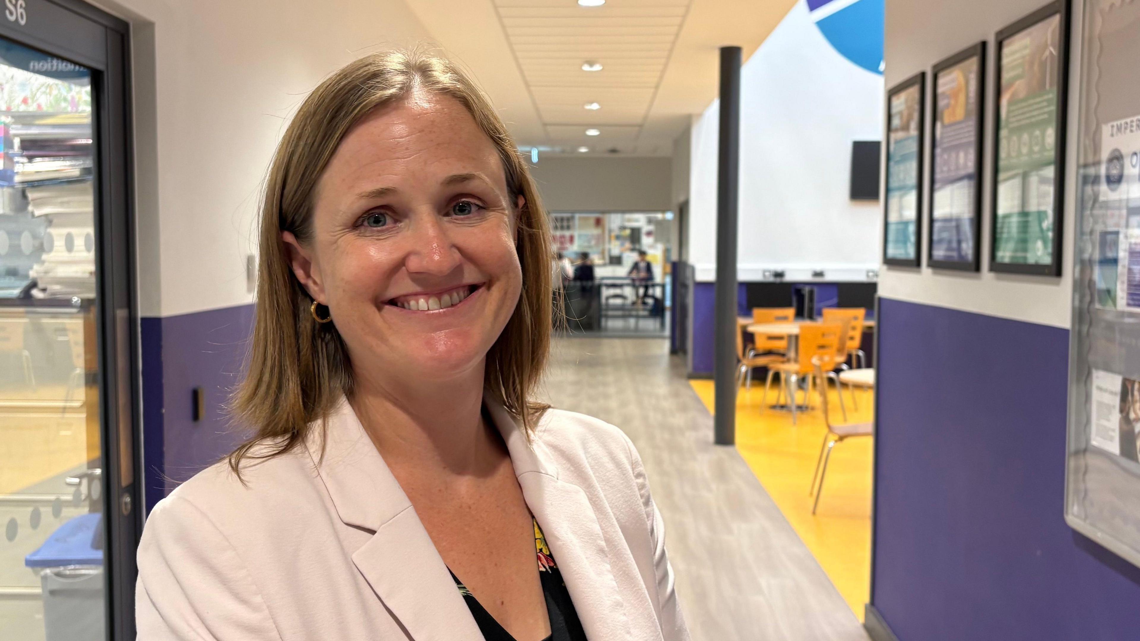 Anna Hewes looking direct to camera. She is standing in a corridor inside Prince William Academy, where the walls are painted purple lower down and white above. In the background are tables and chairs, with a glass-fronted classroom in the further distance. Anna is wearing a white jacket over a dark-coloured dress. She is fully smiling and has dark blonde shoulder-length hair.