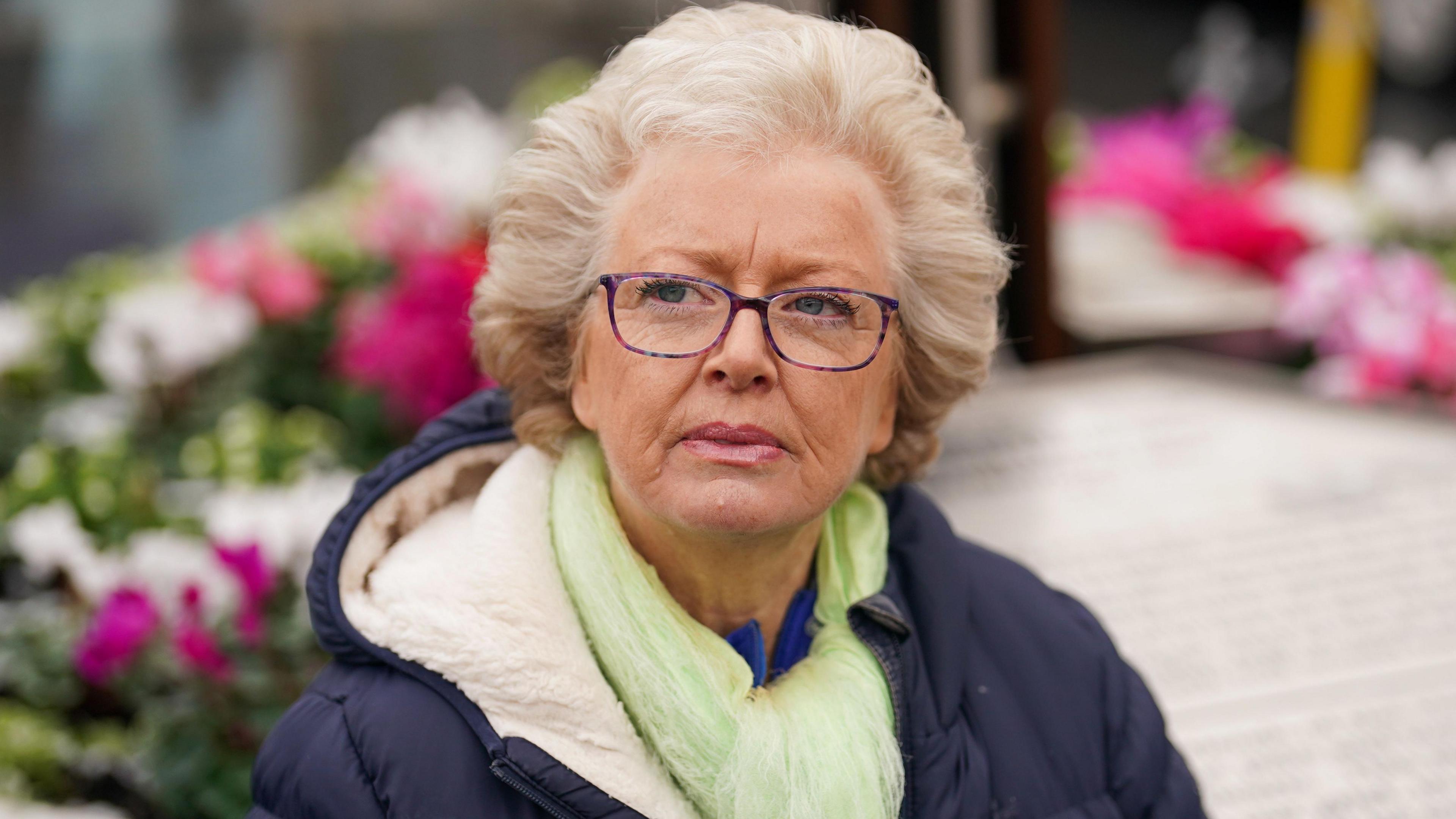 A fair-haired woman stands in front of floral tributes. She is wearing purple-framed glasses, a green scarf and blue coat.