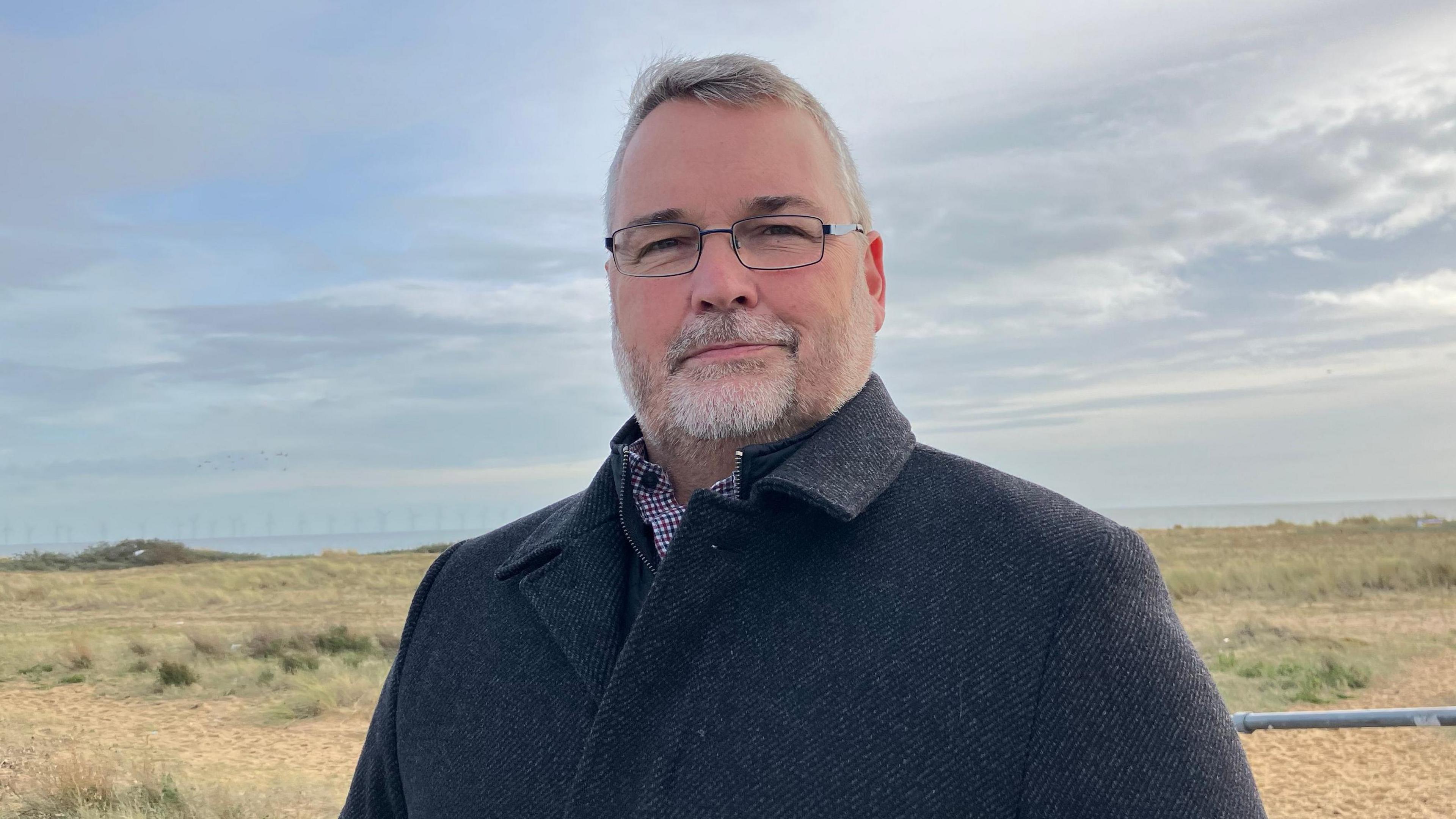 Mark Stephenson wearing a black coat smiling at the camera. He is wearing small frame glasses and a black coat. Behind him is a beach.