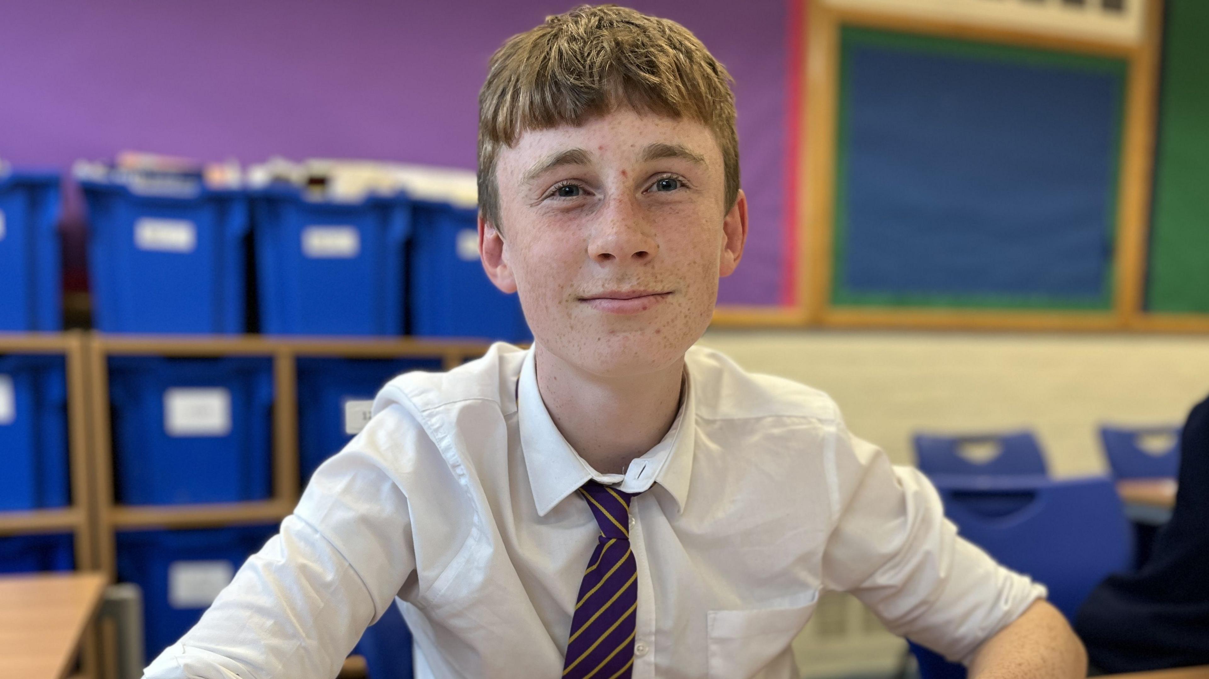 15-year-old Rowan sat at a school desk, smiling, wearing a white shirt and school tie