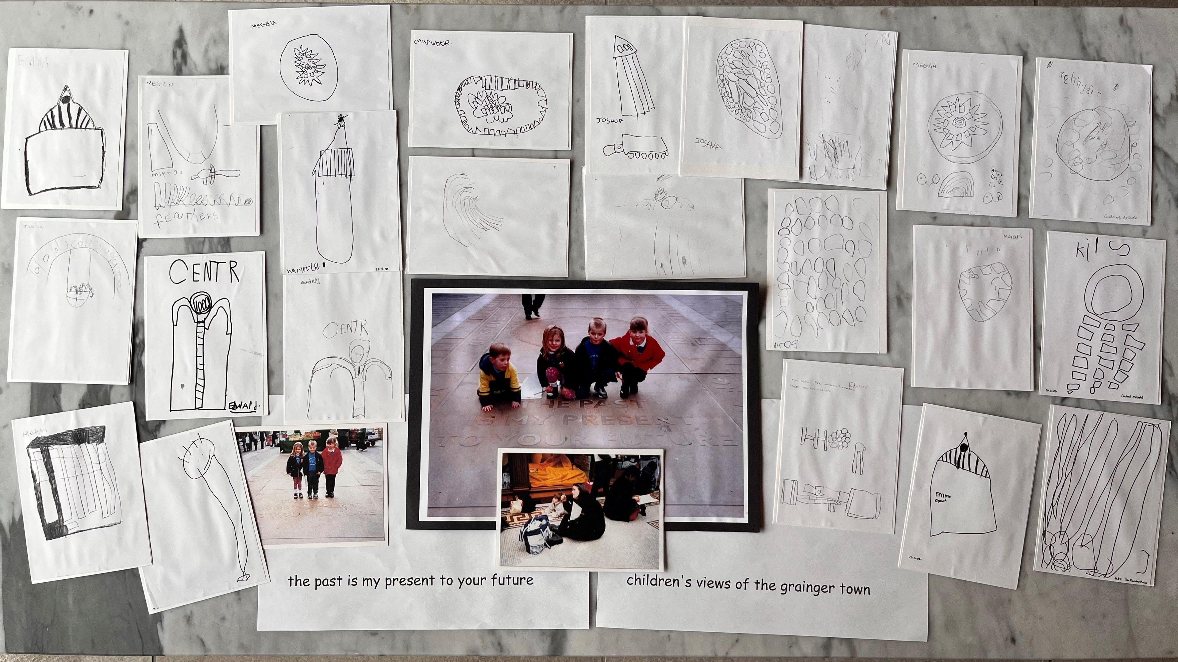 Various bits of white paper with drawings of buildings on them on a table. In the centre is a photograph of a group of school children crouched down on pavement.