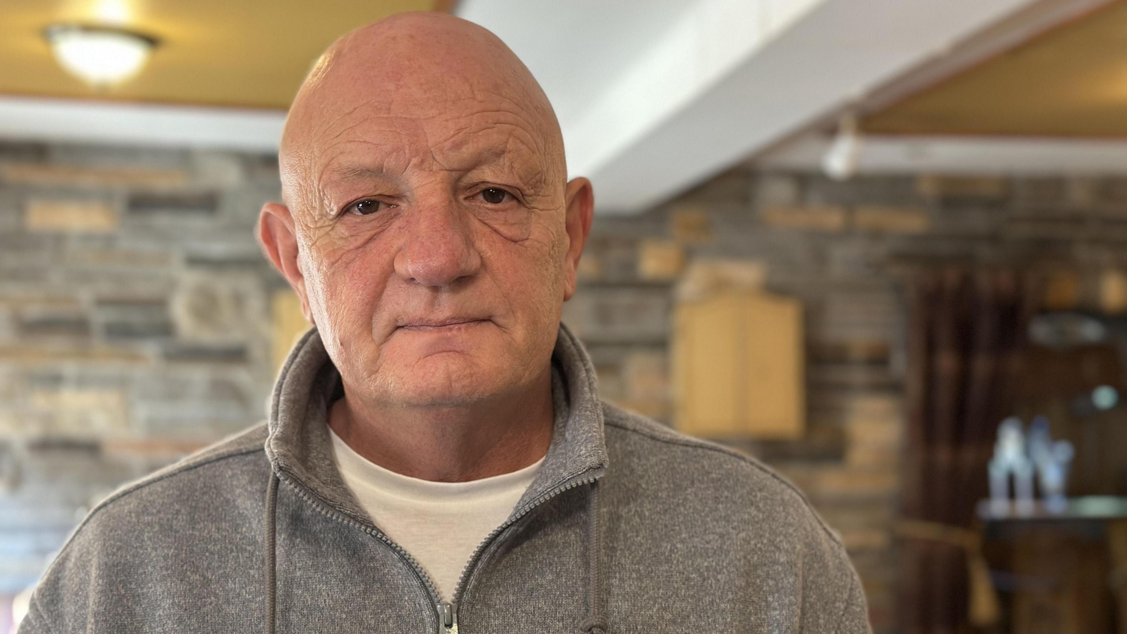 Terence McNaughton is bald with brown eyes. He is wearing a grey zipped top and a white T-shirt underneath. The background is blurred but there is a brick wall and a small wooden cabinet on it.