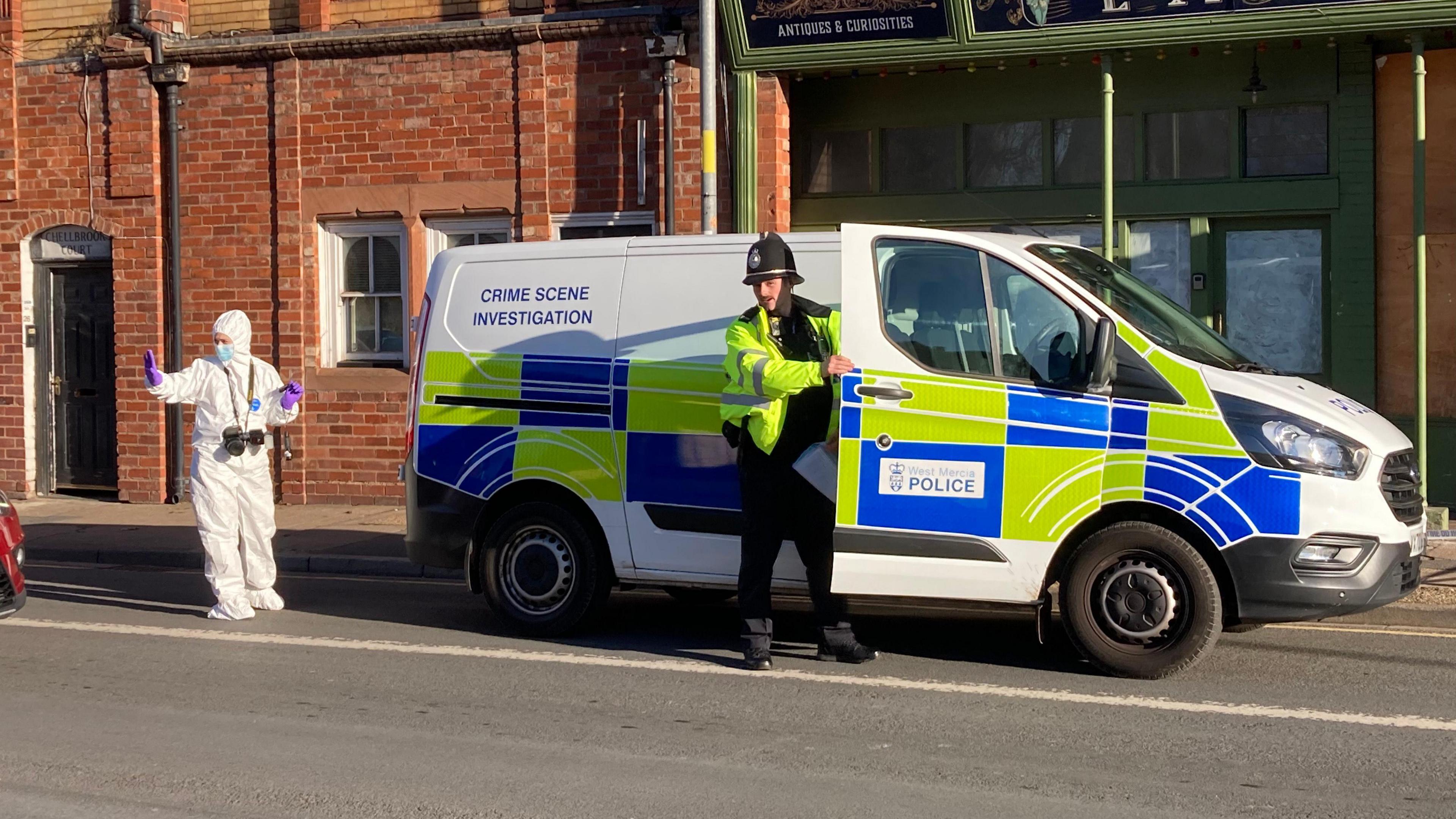 A police officer wearing a bright yellow police coat and black hat opens the door to a white police van with words CRIME SCENE INVESTIGATION on it, parked outside a shop on a road. A second person wears a white forensic suit with purple gloves and puts their hand up to someone out of shot. 