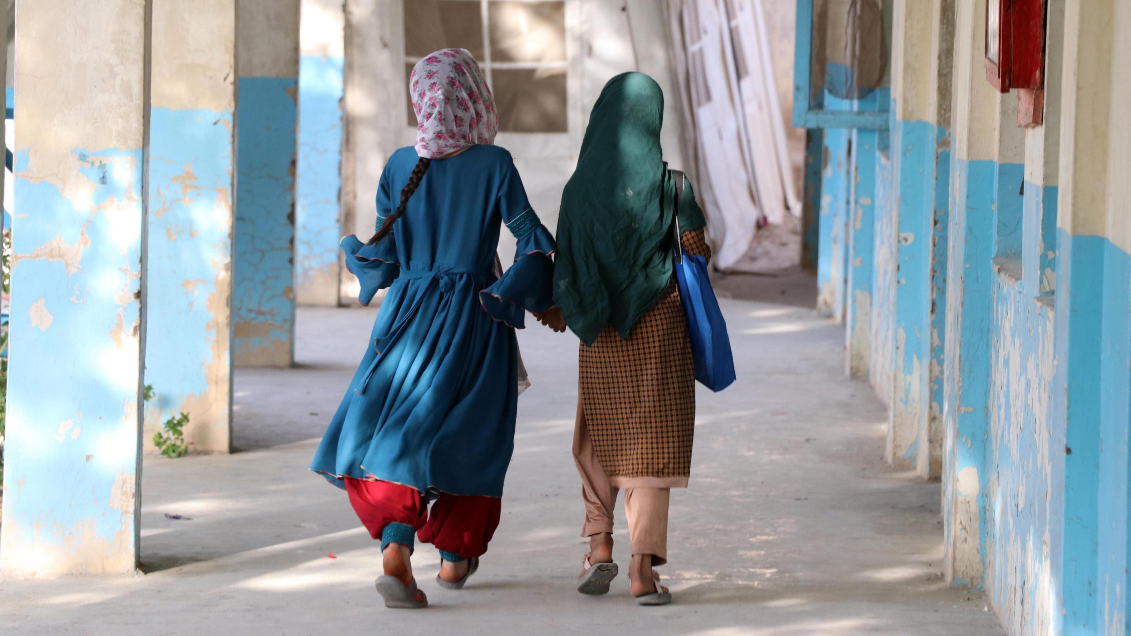 Two Afghan girls walk down an outside corridor with blue and white walls, their hair covered. They wear brightly-coloured clothes, and the sun is shining