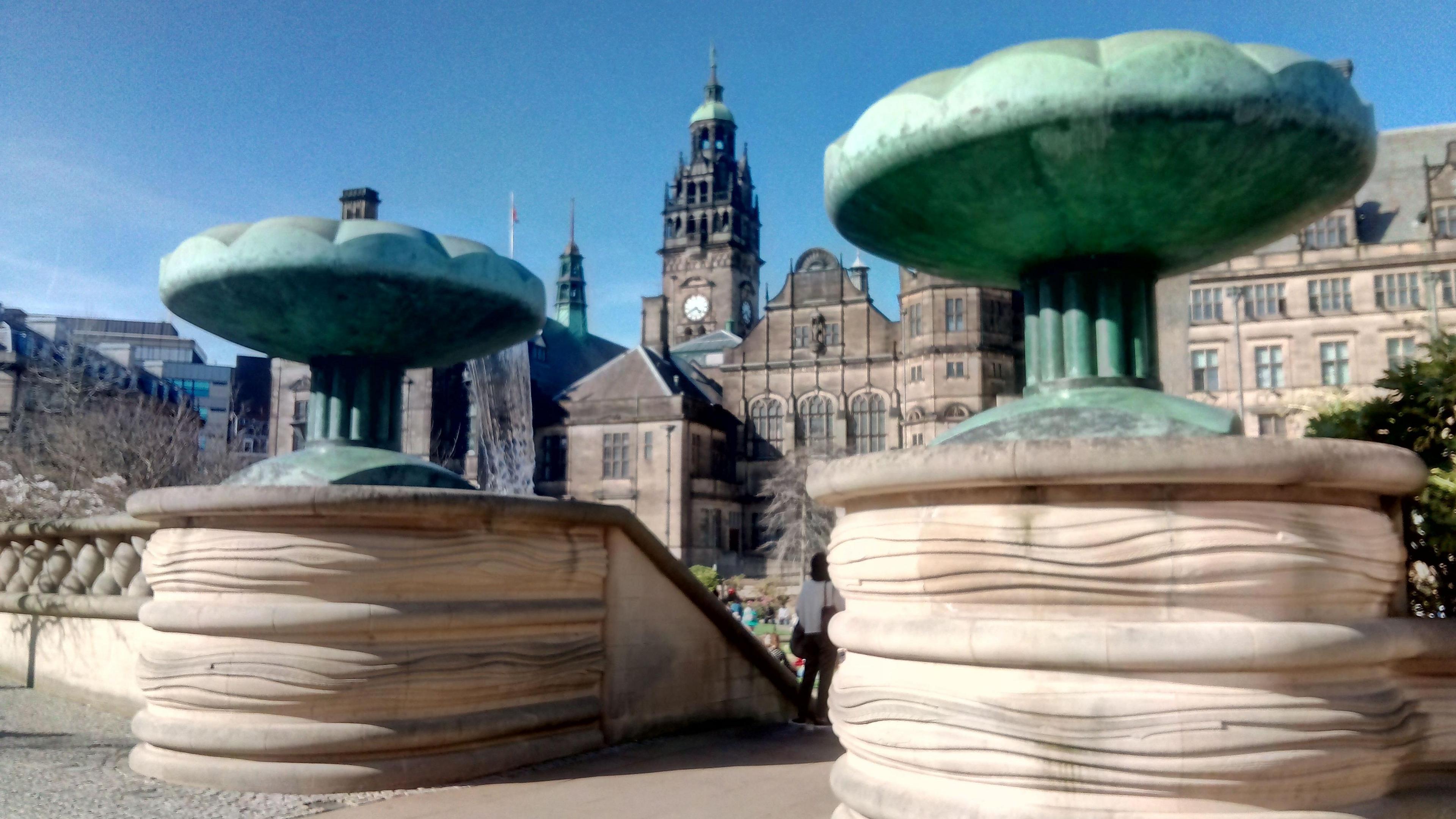 There are two large green stone structures at the top of steps leading to a paved area and fountains in Sheffield Peace Gardens. The town hall is in the background