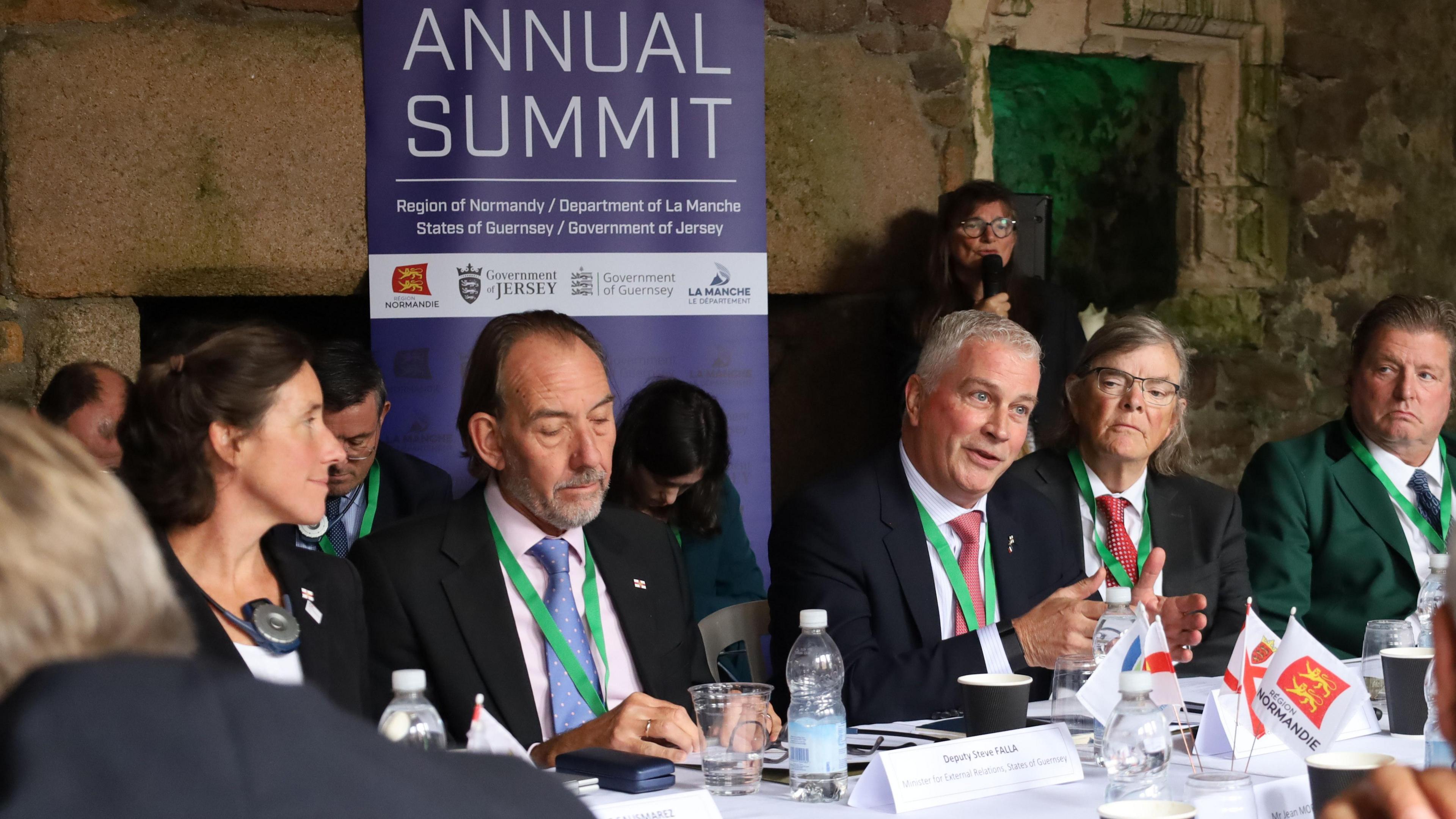 Men and women wearing suits, sit at a table in conversation. Behind them is a purple poster that reads 'annual summit.