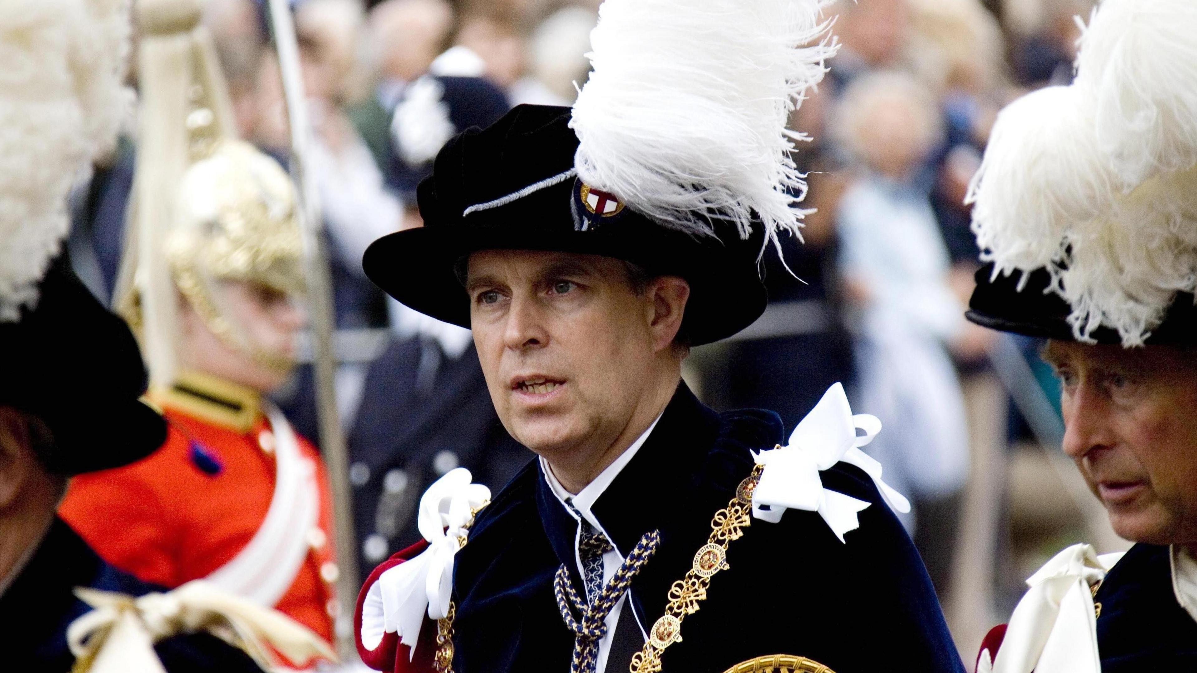 Prince Andrew in the regalia of the Order of the Garter. He is wearing a black jacket and hat with gold chains, white ribbons and a white feather from his cap. The King, wearing the same regalia, can be seen to his left