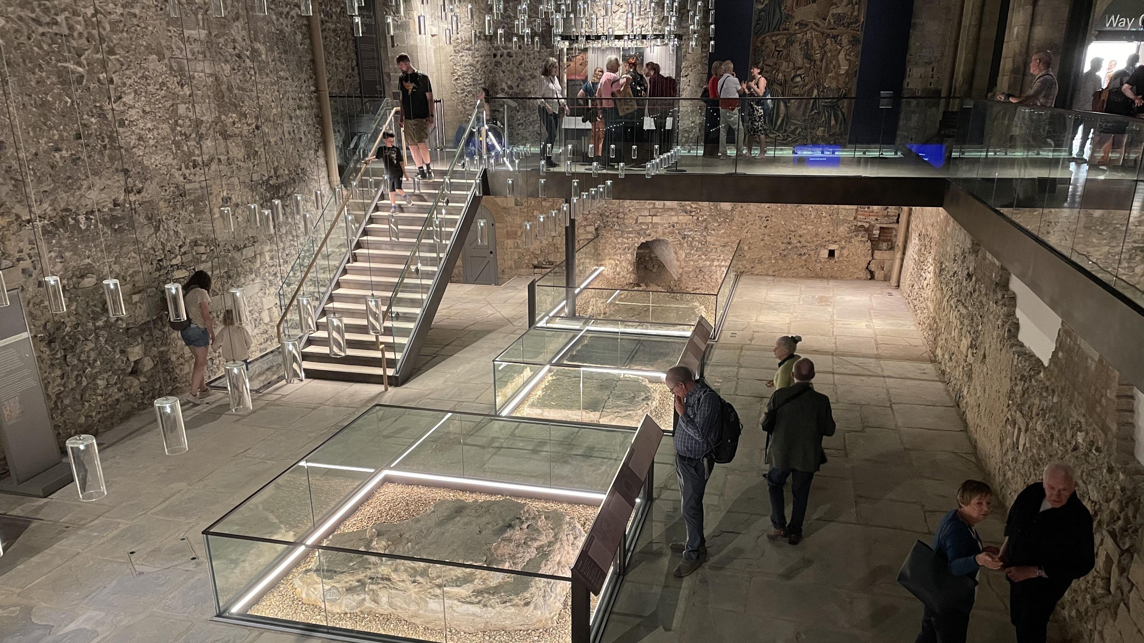 Small groups of people look at glass exhibition spaces showing artefacts in a Norman castle, with stairs to the upper level on the left.