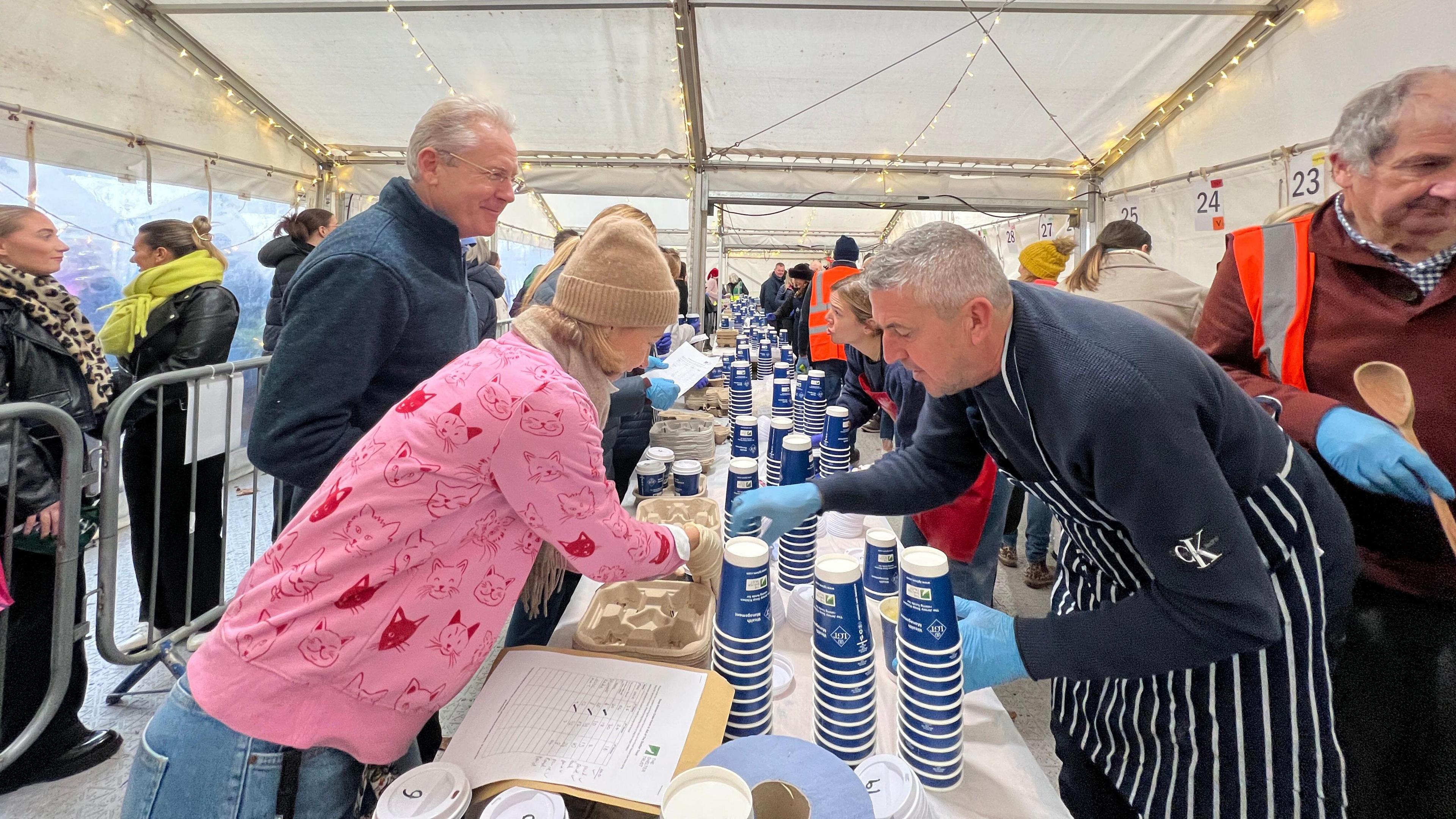 People lean over a line of tables containing soup holders. Those on the right side of the table have plastic gloves and are taking orders from the people on the left who are collecting the soup for customers.