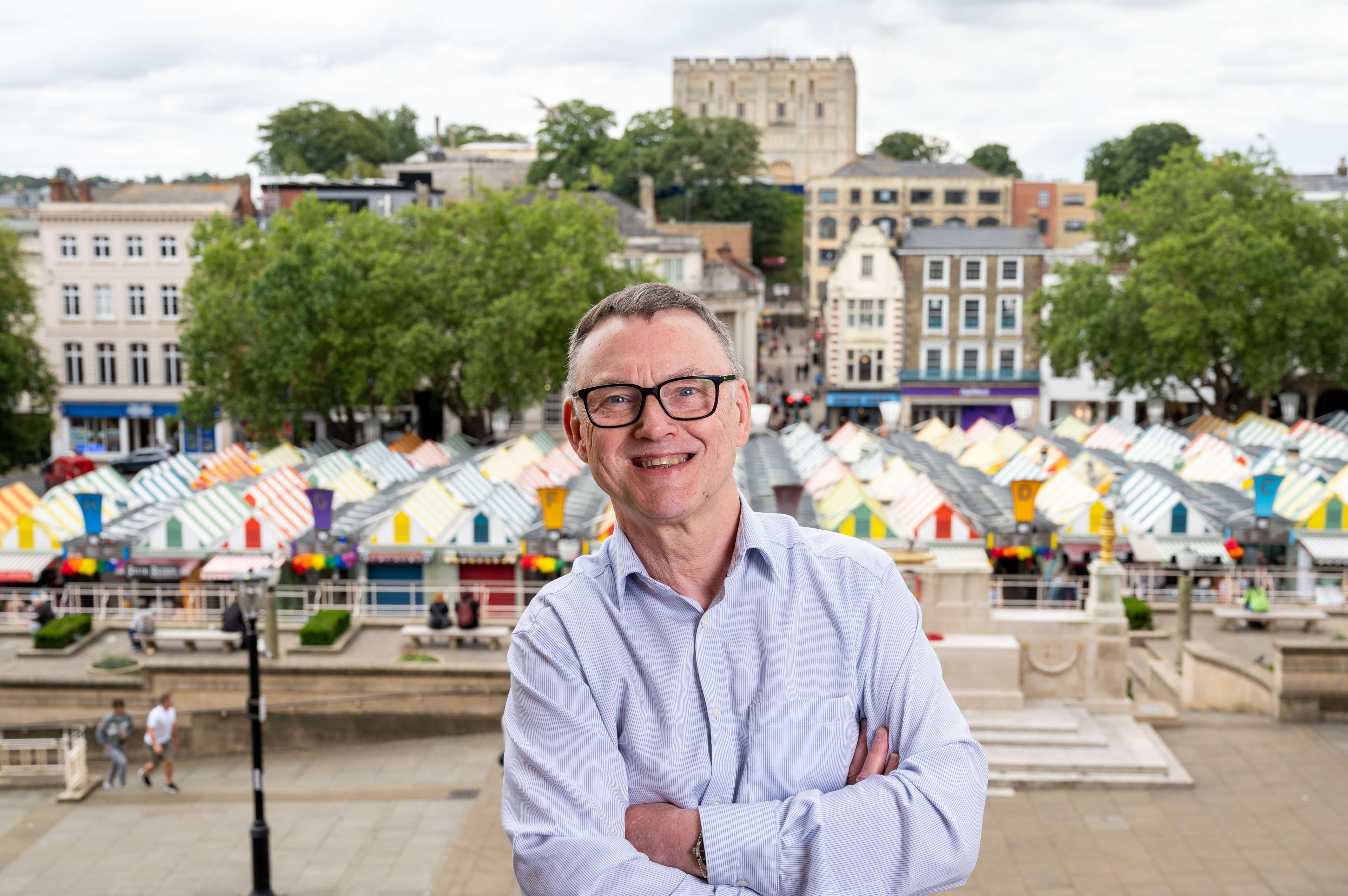 Mike Stonard, wearing a light-coloured shirt and black glasses, smiles at the camera. He has short grey hair. Behind and below him are the brightly-coloured canopies of Norwich Market's stalls. 