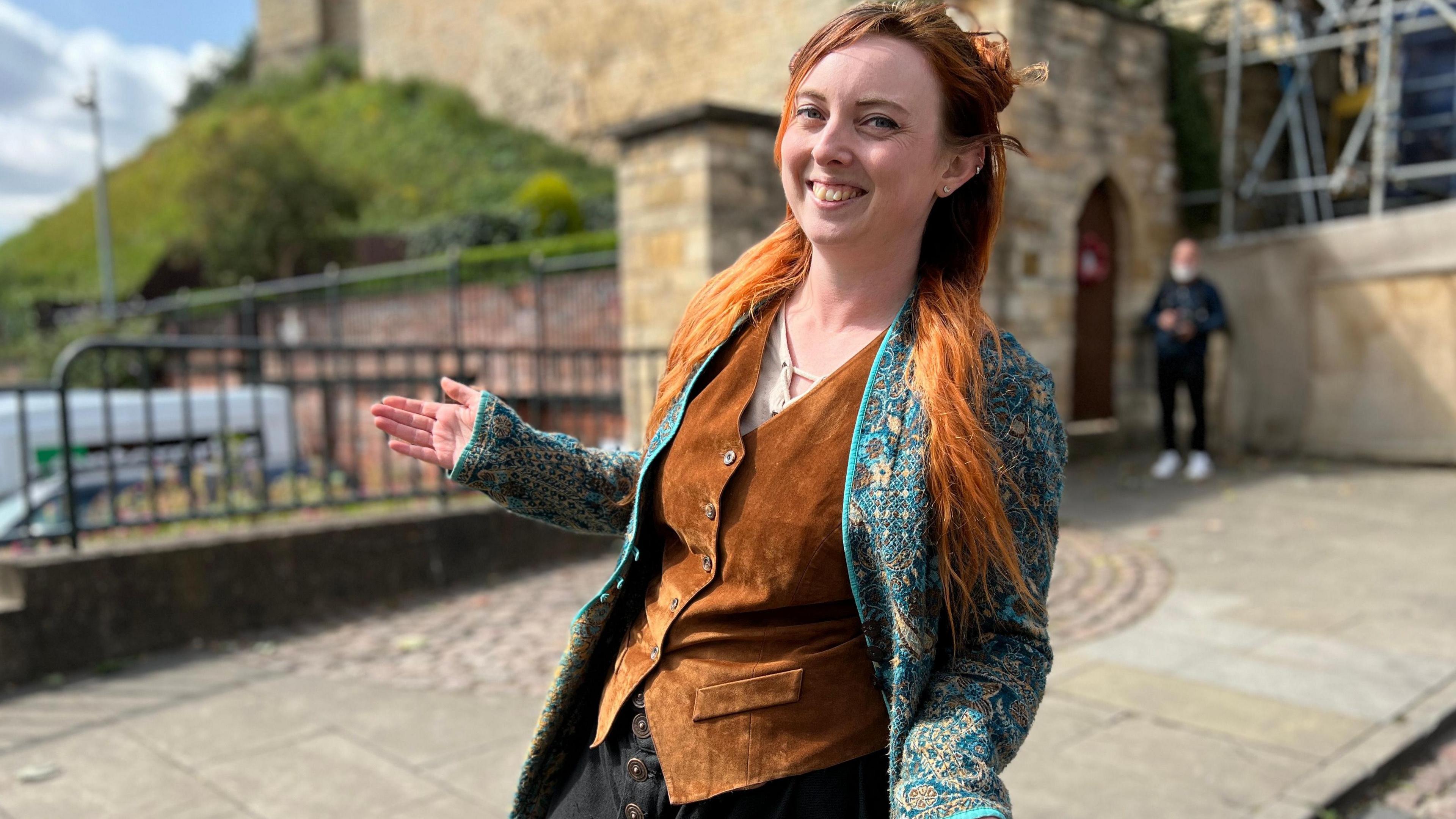 Woman with long red hair with arms out to the side dressed in a brown waistcoat and paisley jacket 