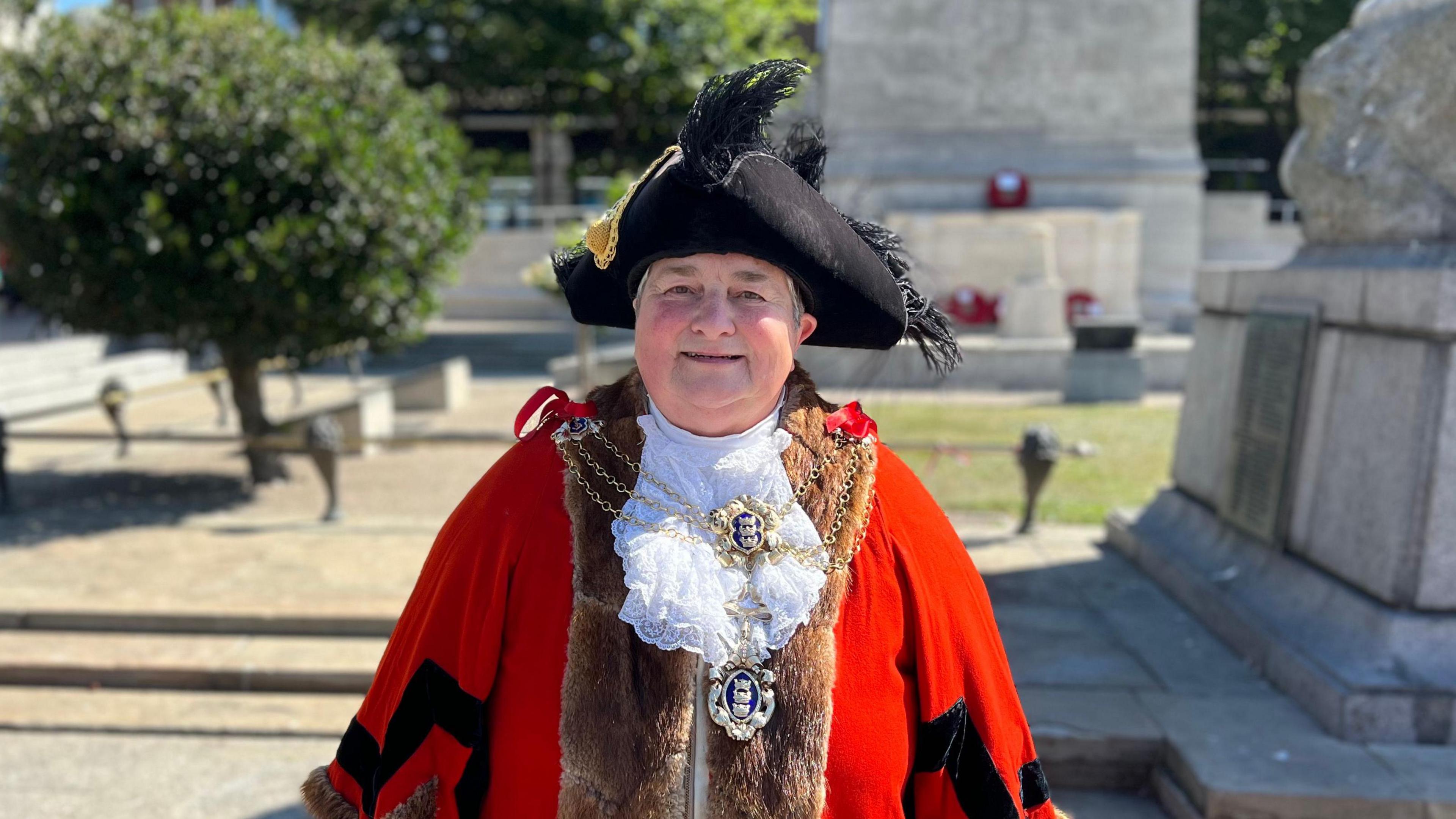 A head and shoulders shot of a woman dressed in ceremonial robes and a tricorn hat. The red robes have a fur trim, and she is also wearing a chain and medallions. She is standing outdoors on a sunny day with a monument and trees behind her.