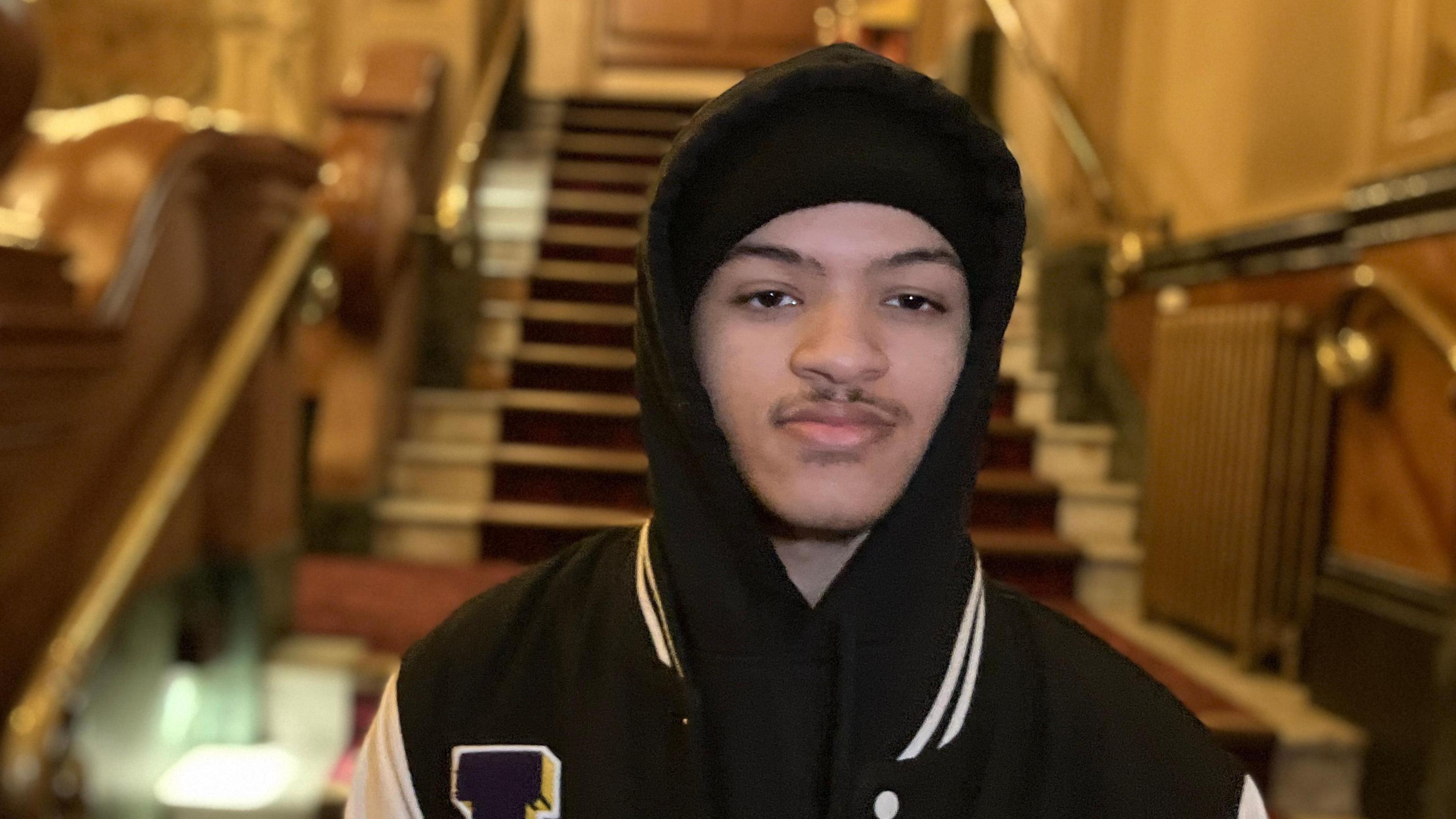 Tryelle Johnson, wearing a black jacket with white lines on the collar and a black hat, is looking straight at the camera. Stairs from the theatre can be seen in the background