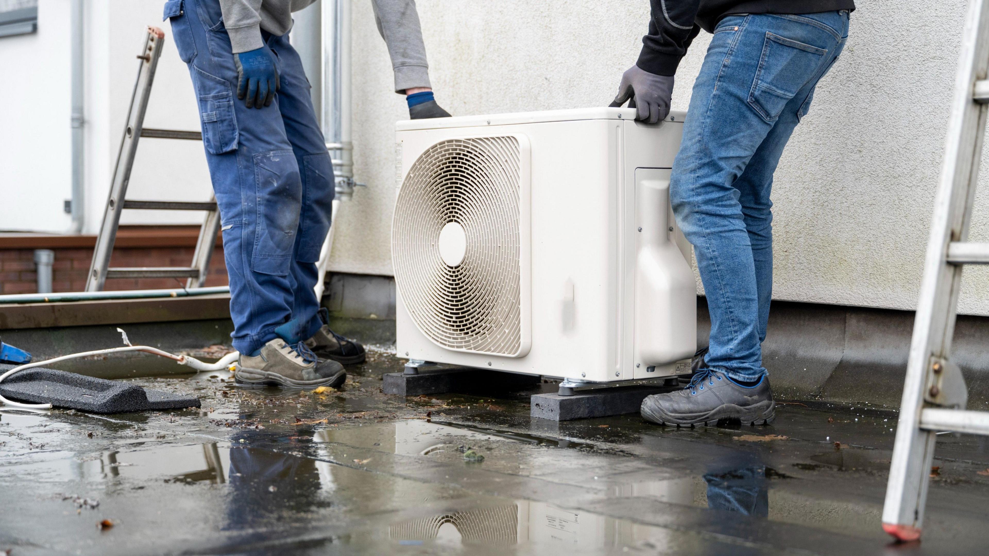 Two construction workers installing a heat pump on a new property, each one holding an end.