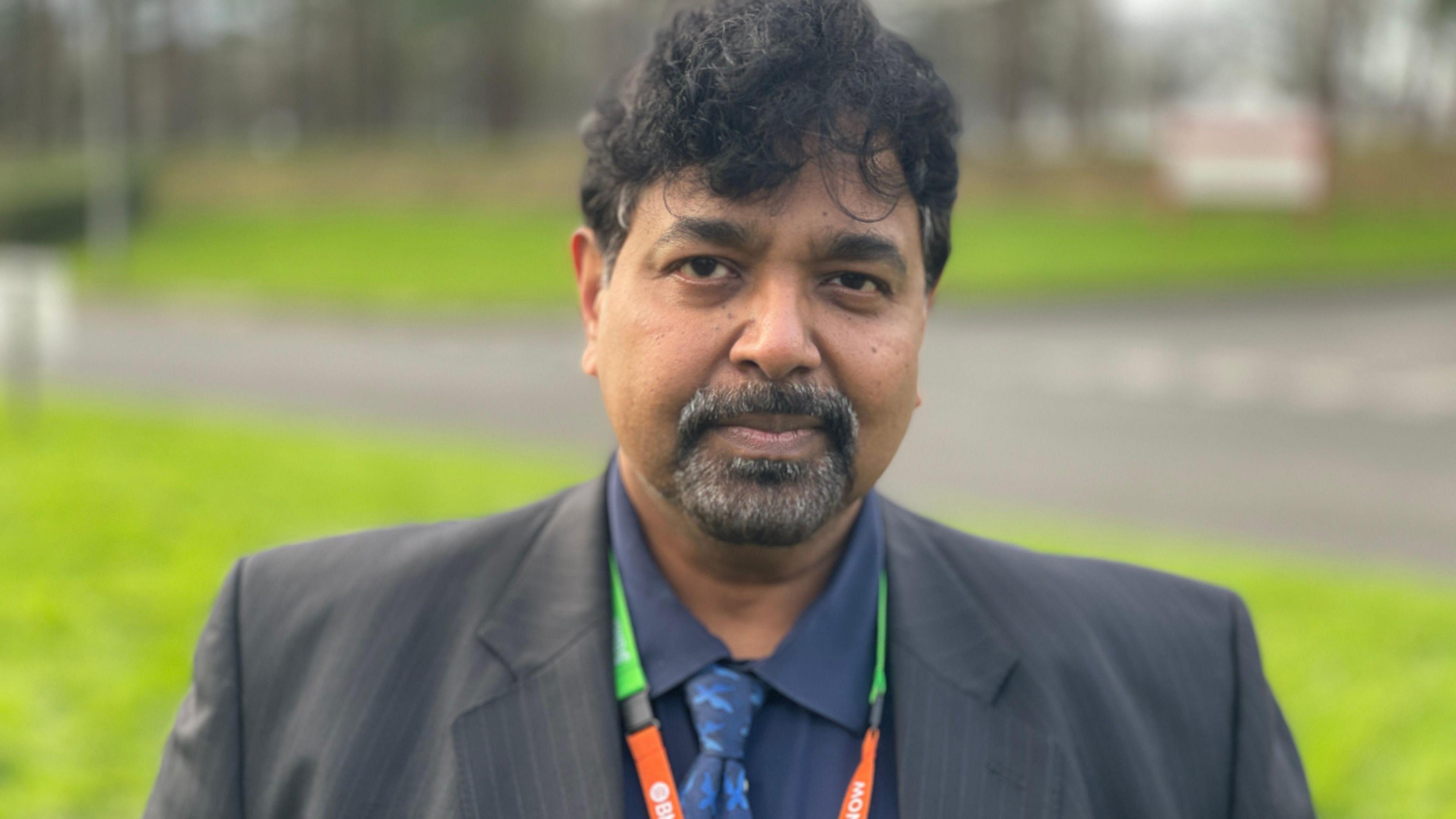 Prakash Thiagarajan standing on a grass embankment wearing a grey suit jacket and blue shirt and ties. He has dark hair and a goatee beard and an orange and green lanyard is hanging around his neck.