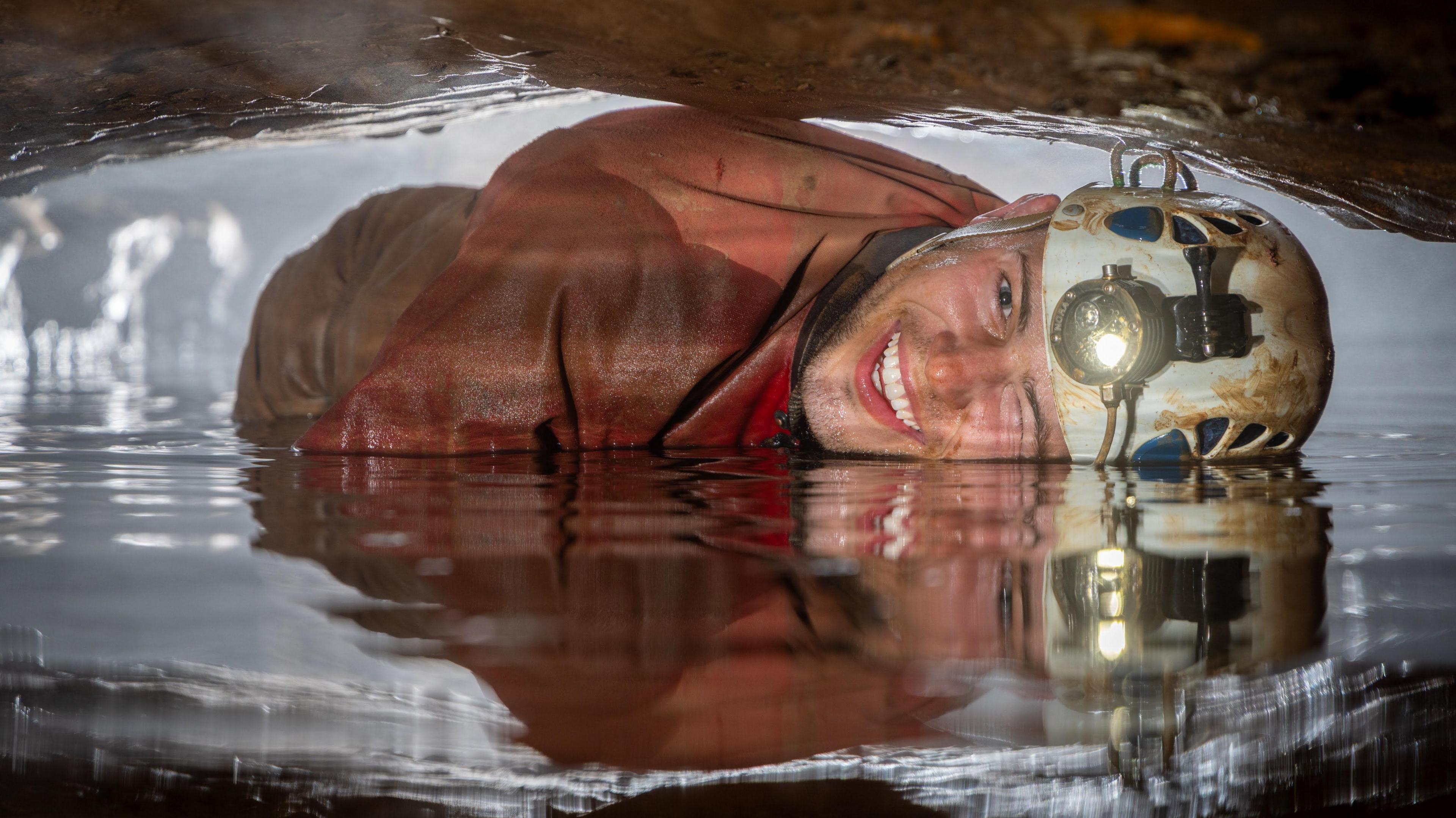 A male caver, wearing an orange waterproof outfit, leans to the side as he passes through a narrow section underground. Part of his face is in the water as he smiles at the camera. He is wearing a white safety helmet with a round head lamp on the front