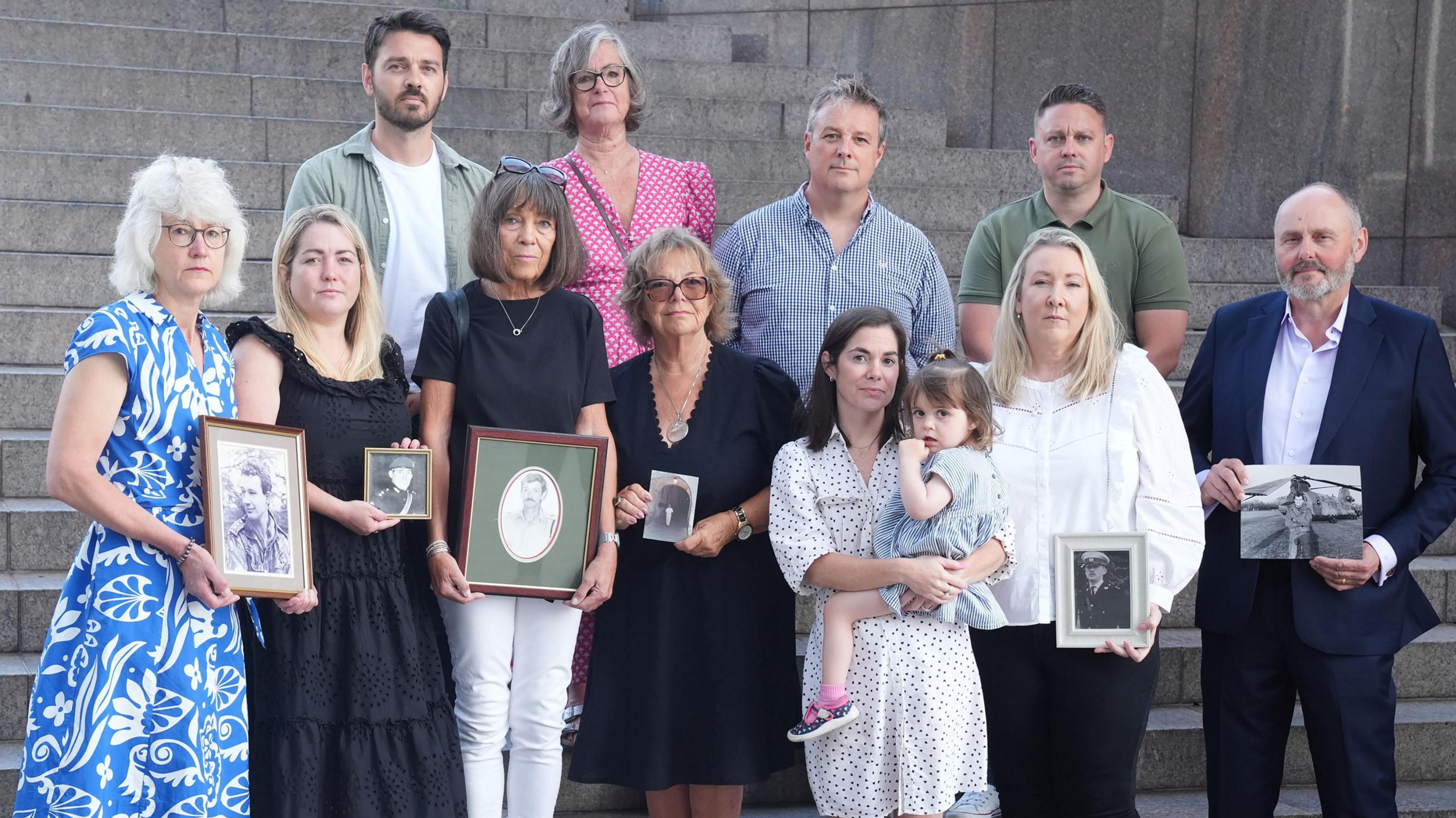 Eleven members of the Chinook Justice Campaign. Some of the group are holding photographs of their loved ones. Within the picture is three members of the Tobias family including Gaynor Tobias who is the third person standing in the front and is wearing a black T-shirt with white trousers and black trainers and she is holding a photograph. On the back row, is Andy Tobias, who is wearing a T-shirt with a khaki shirt over the top and at the end of the back row is Matt Tobias who is wearing a khaki coloured T-shirt. All members of the group are standing looking directly at the camera.
