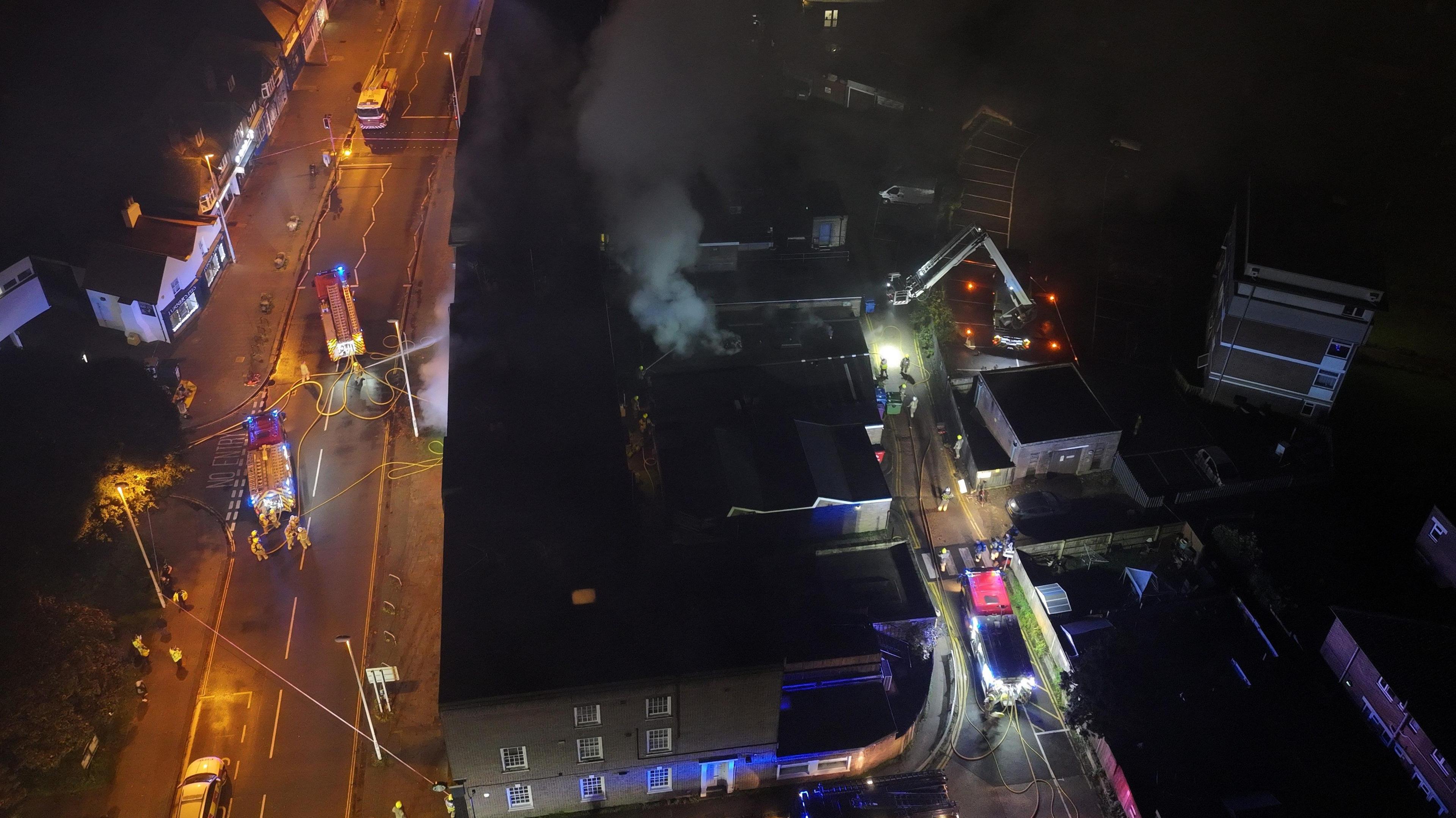 An aerial view of smoke billowing from a property. Fire engines line the street outside. 