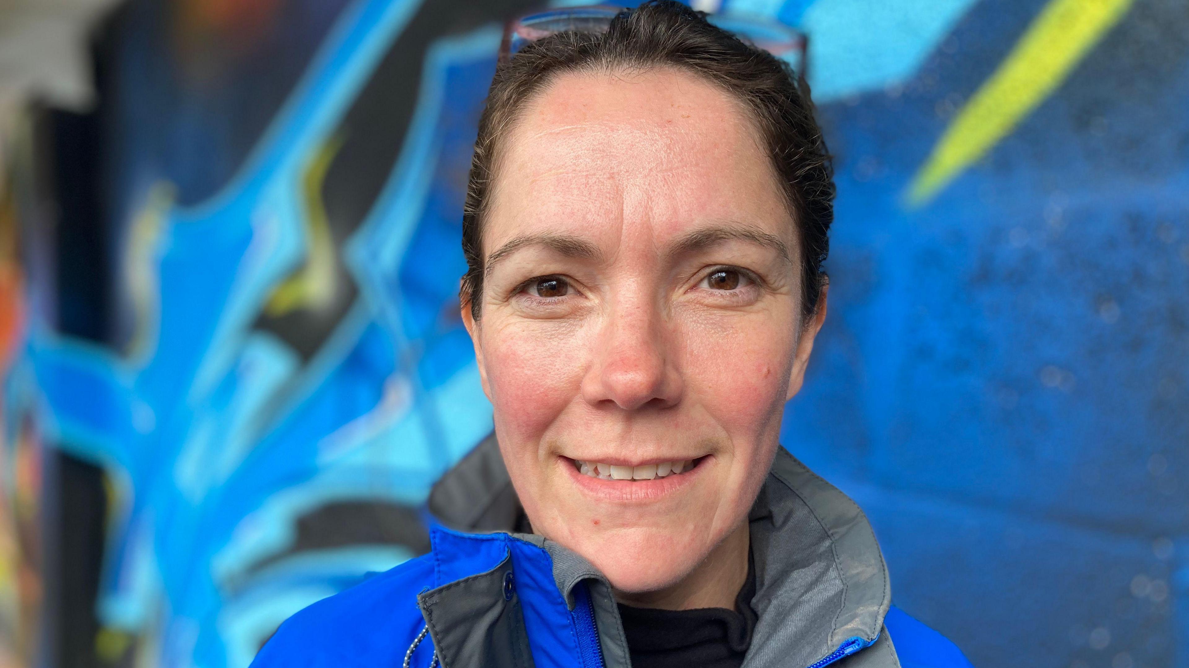 Head shot of a woman with brown hair and wearing a rain jacket standing in front of a wall covered in a blue coloured mural