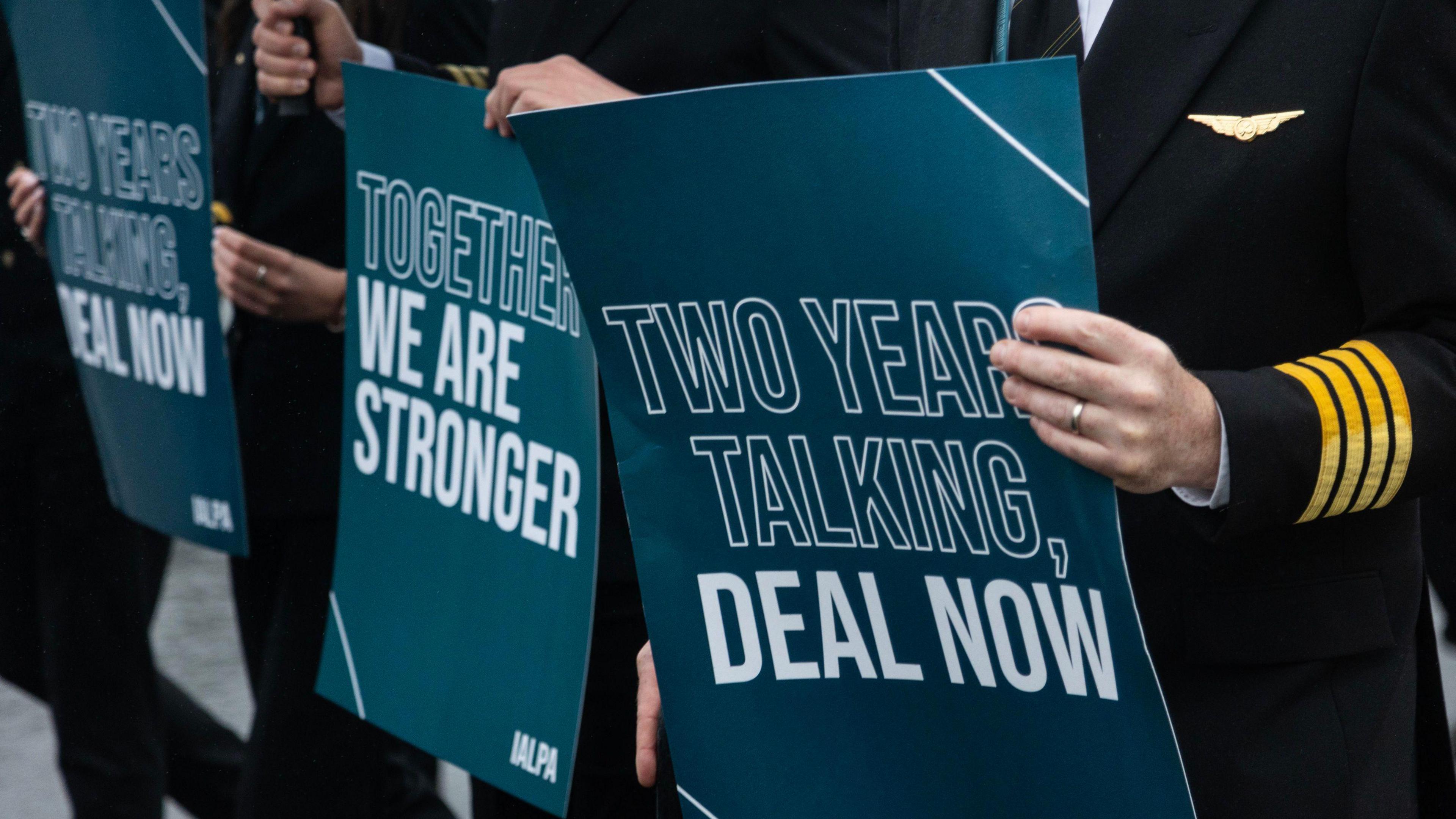 Pilots on strike holding signs that read 'together we are stronger' and 'two years talking, deal now'. The pilots are in uniform (black blazers and trousers with yellow stripes on the cuff and a plane gold badge on the lapel.) The signs are green/blue with white writing.