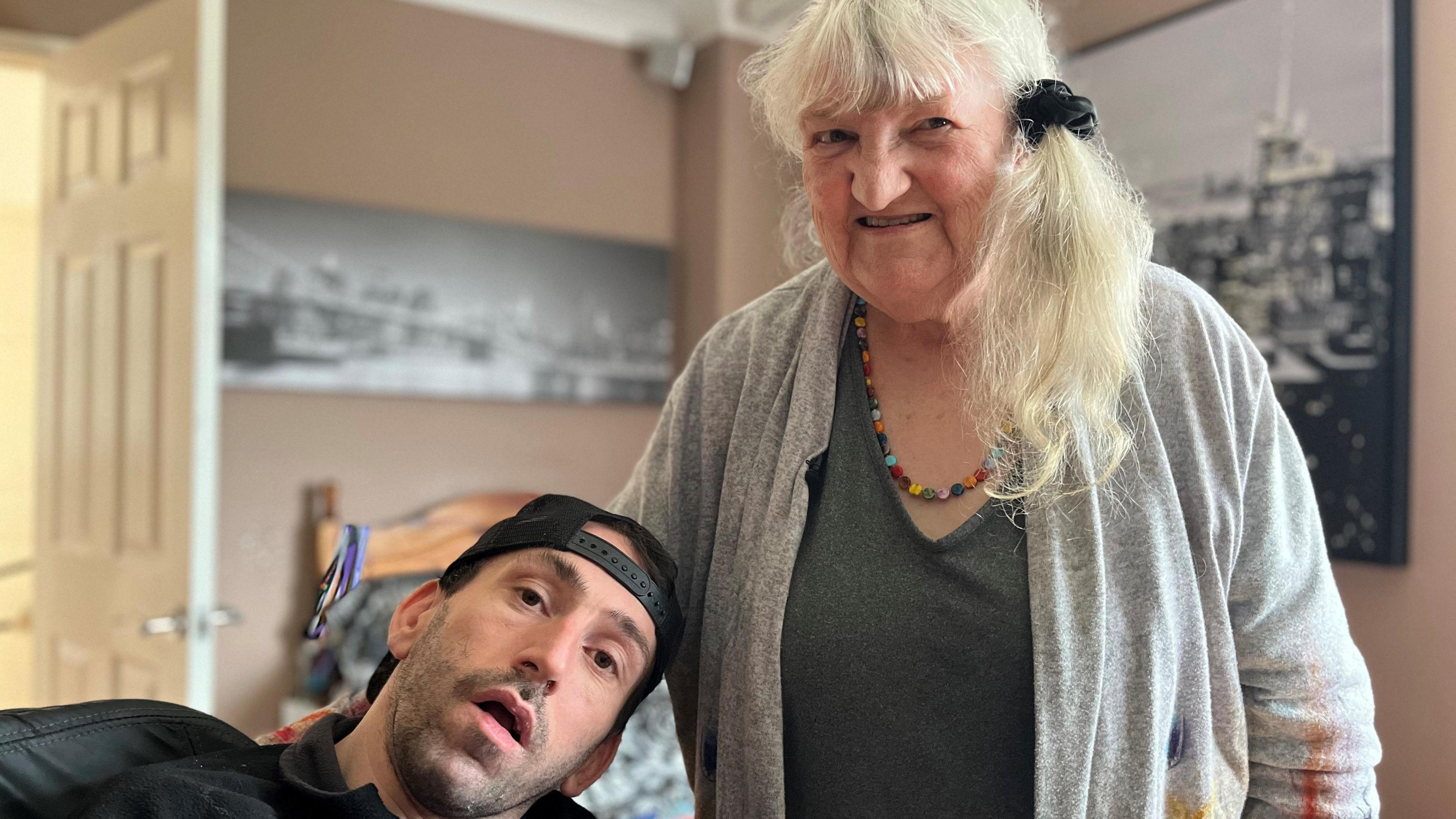 Gareth Stafford and his mother Gill. Gareth is sat in his electric wheelchair, and is wearing a back-to-front baseball cap. His mother Gill has her arm around him and is smiling at the camera.