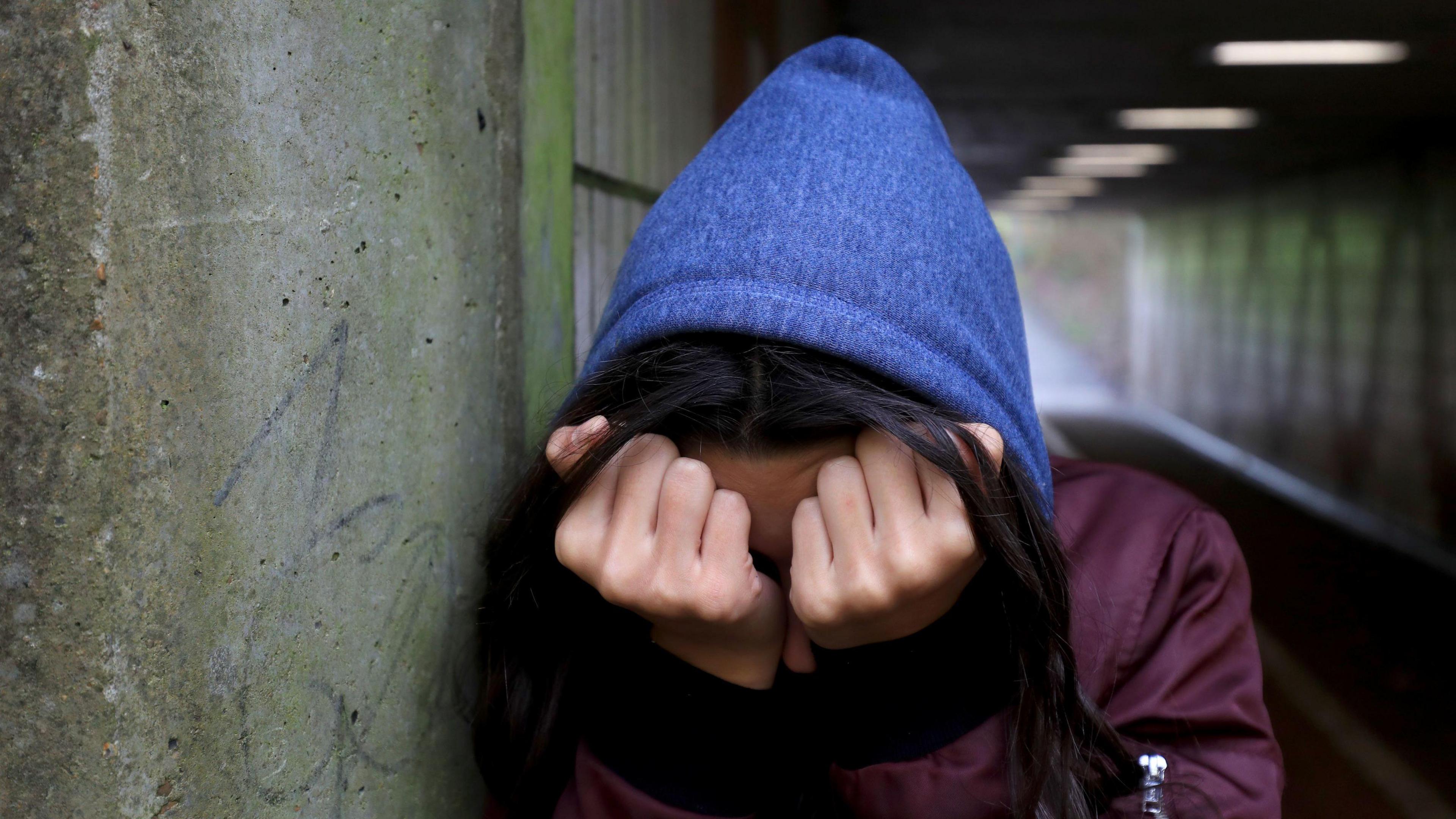 A young women holds her head in her hands, covering her face. She is wearing a blue hoodie and standing in a dark underpass.