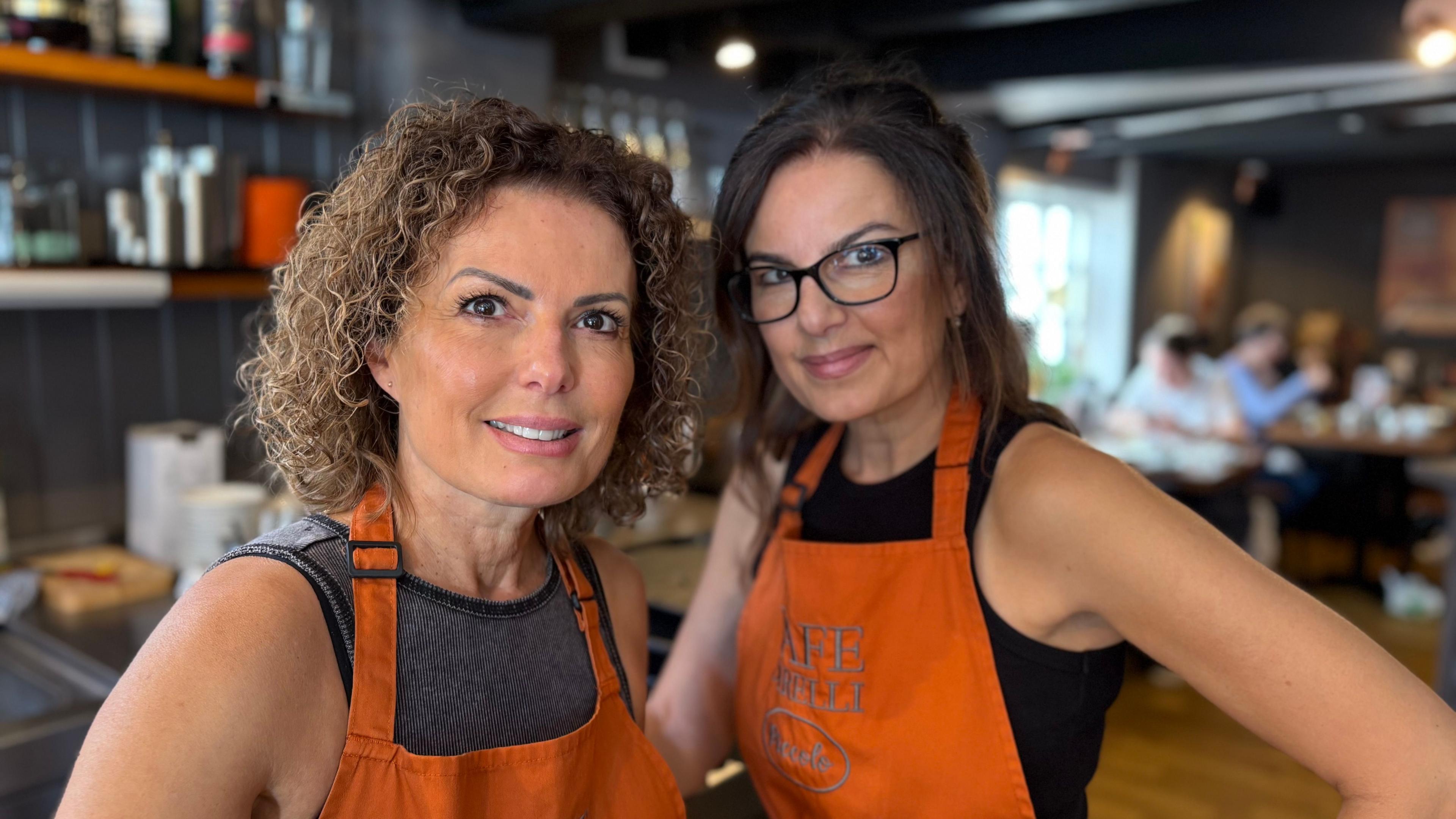 Serafina Zambito with long straight brown hair and glasses, and Jo Garvin with curly brown hair, both wearing orange aprons standing in a cafe with items on shelves behind them to the left and customers sitting at tables in the background.