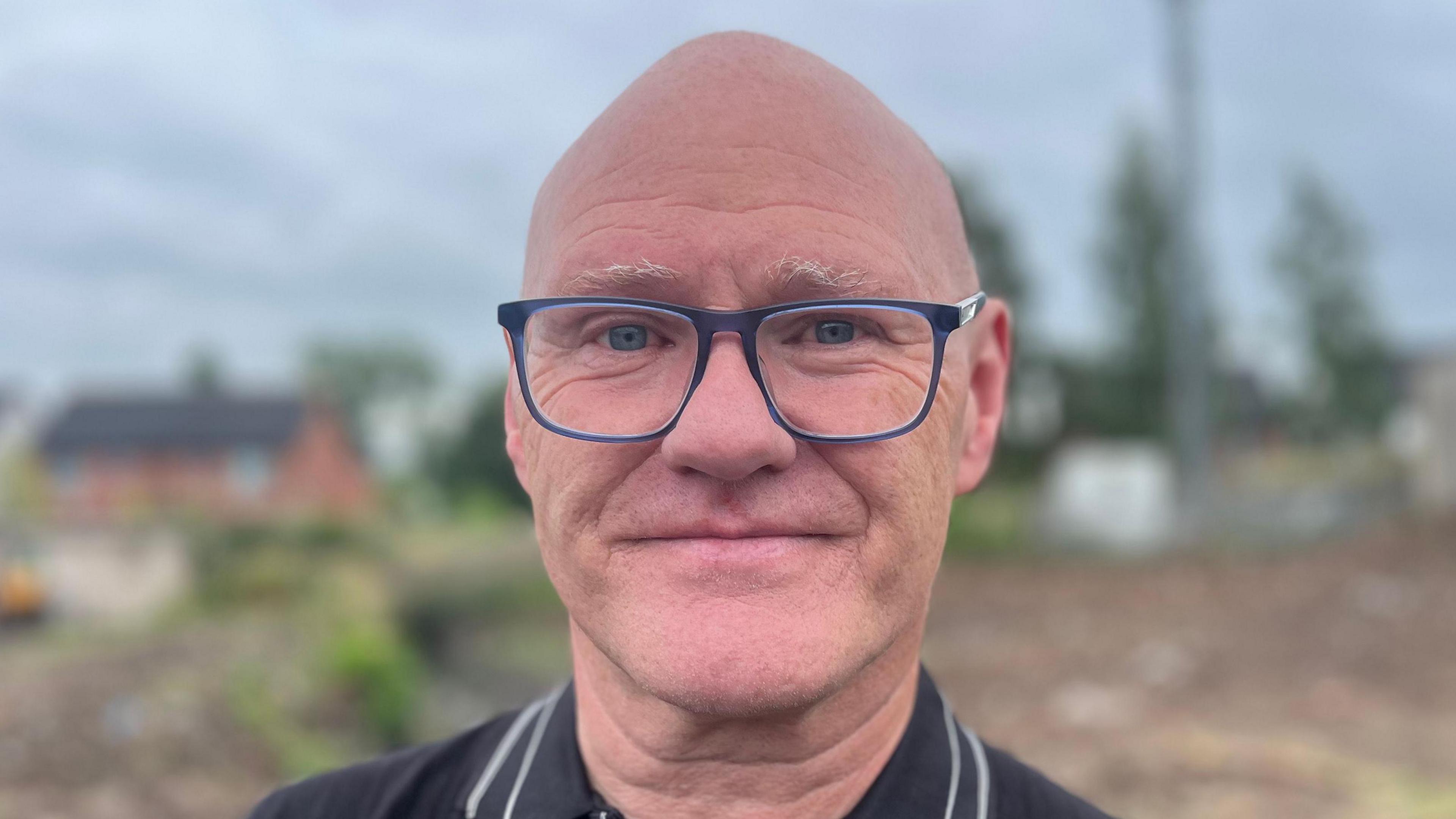 Paul Maskey is standing near a 5G mast and is looking face on. He has a bald head and wearing blue glasses and a black polo shirt with white lines on the collar. 