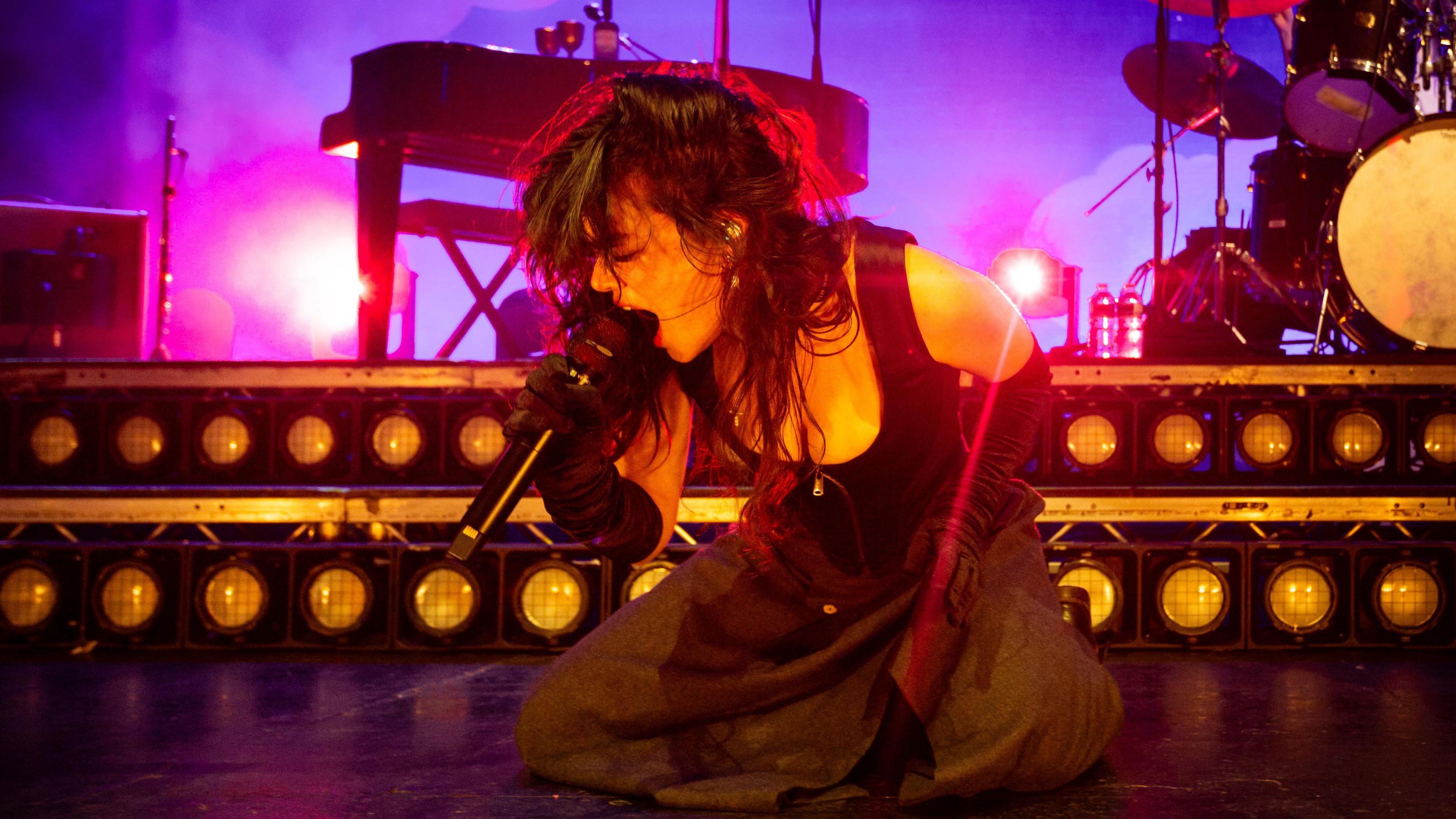 Abigail Morris of The Last Dinner Party performs at O2 Academy Leeds on 24 September. Abigail kneels on stage as she sings into a microphone, her long dark hair worn loose. She wears a black skirt, long black gloves and corset-style top. Behind her there's a piano and drum kit, the stage is lit purple behind her. 