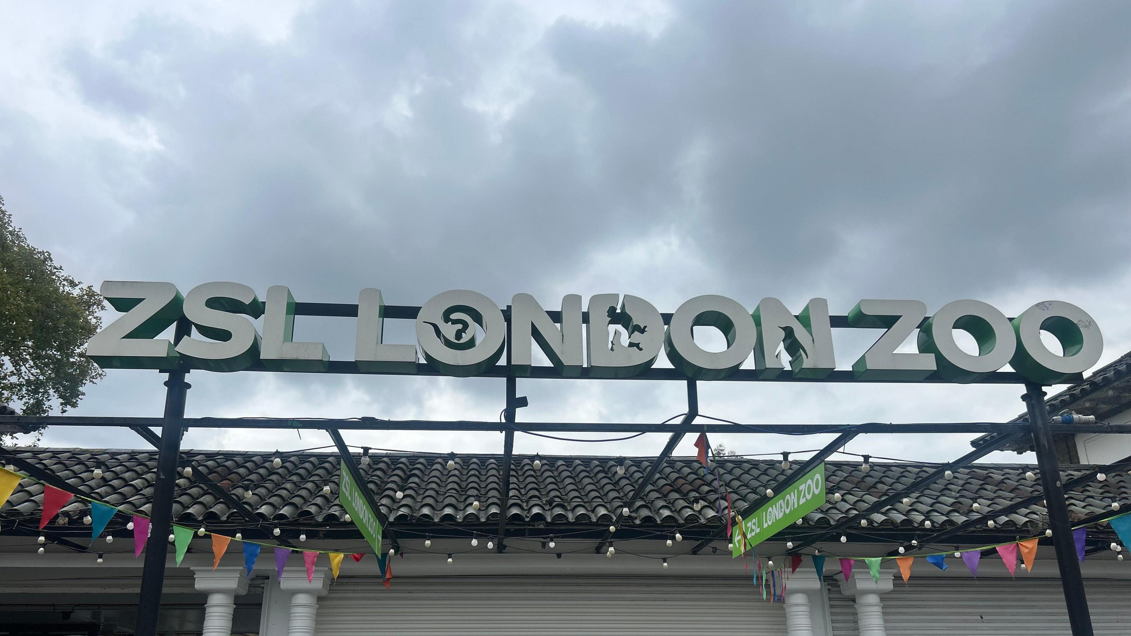 A sign for London Zoo at entrance of the zoo against a background of grey clouds. 