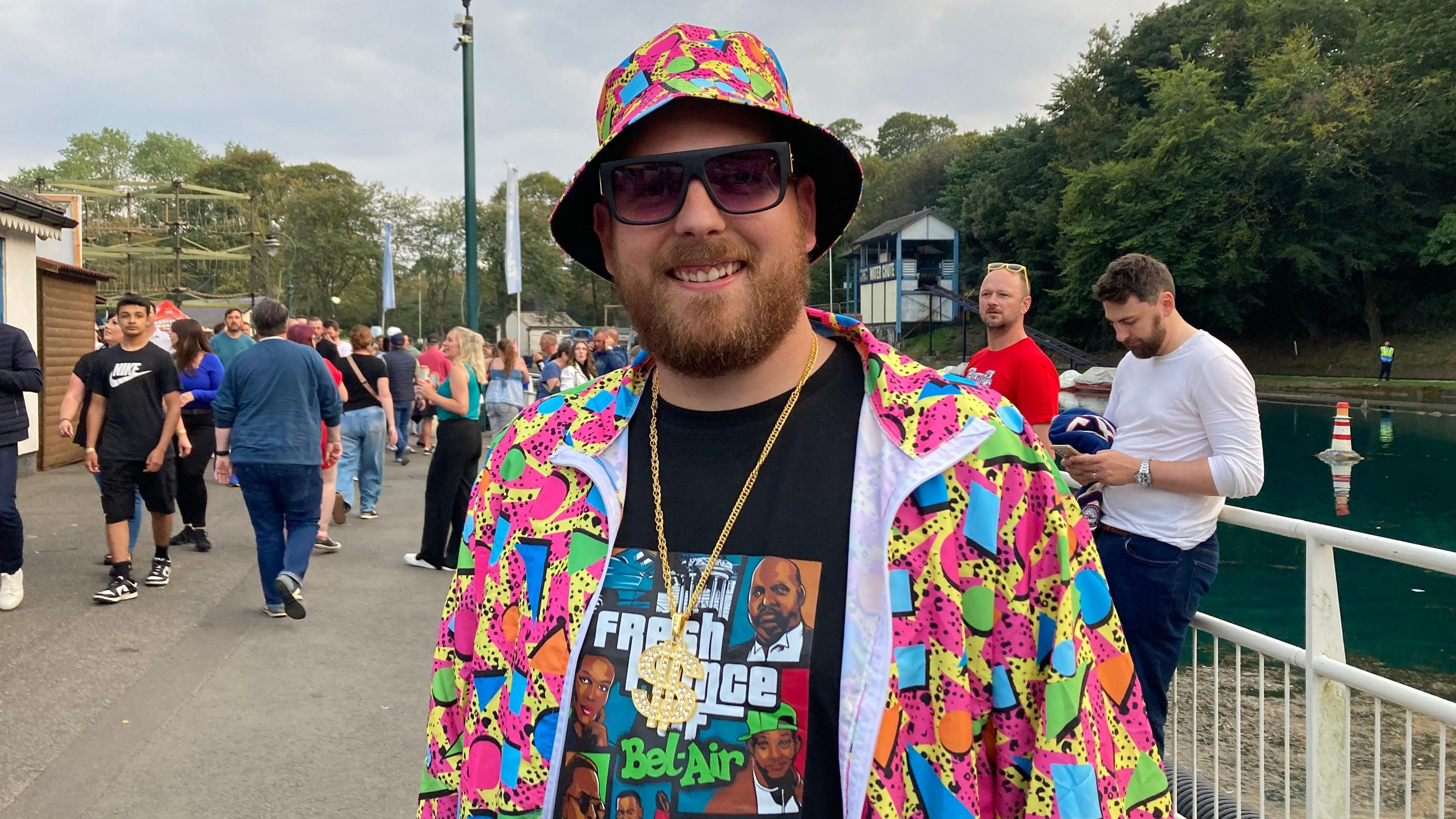 Jordan Taylor is pictured in a brightly-coloured shirt which is unbuttoned. Underneath the shirt is a Fresh Prince of Bell-Air T-shirt. He is also wearing a brightly-coloured bucket hat, a chunky gold chain and sunglasses and he is standing in front of a lake. There are people milling about behind him.