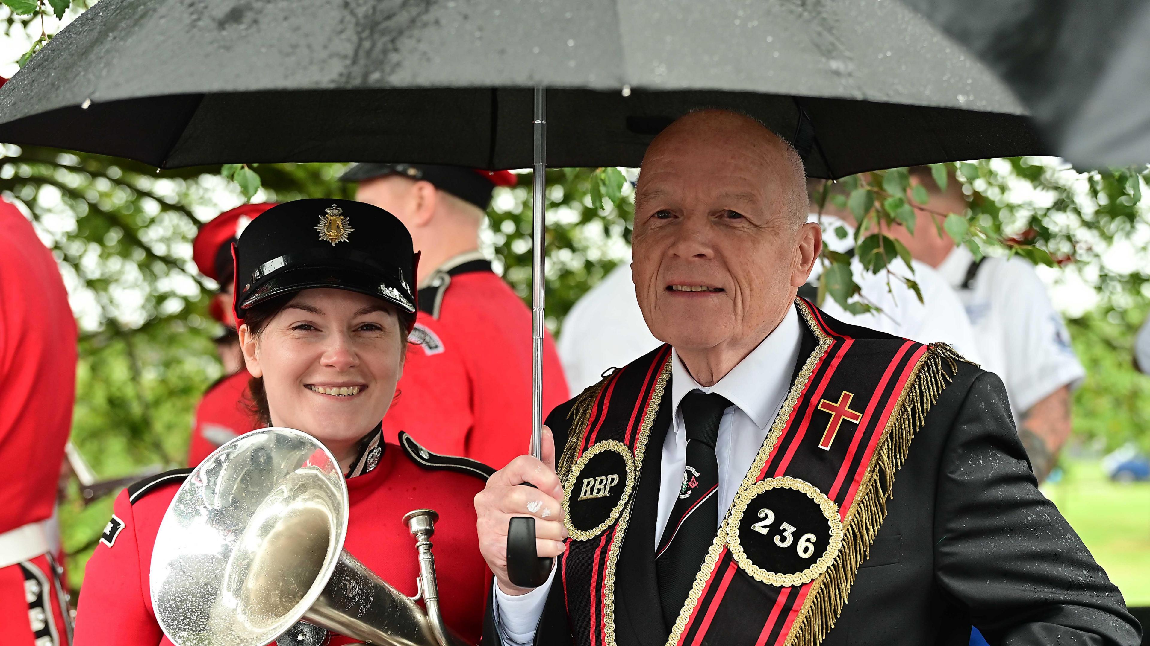 A woman in a red suit with a black peaked cap is holding a silver tuba. A man stands beside her in a black suit with a black sash with red trim. He holds an umbrella.