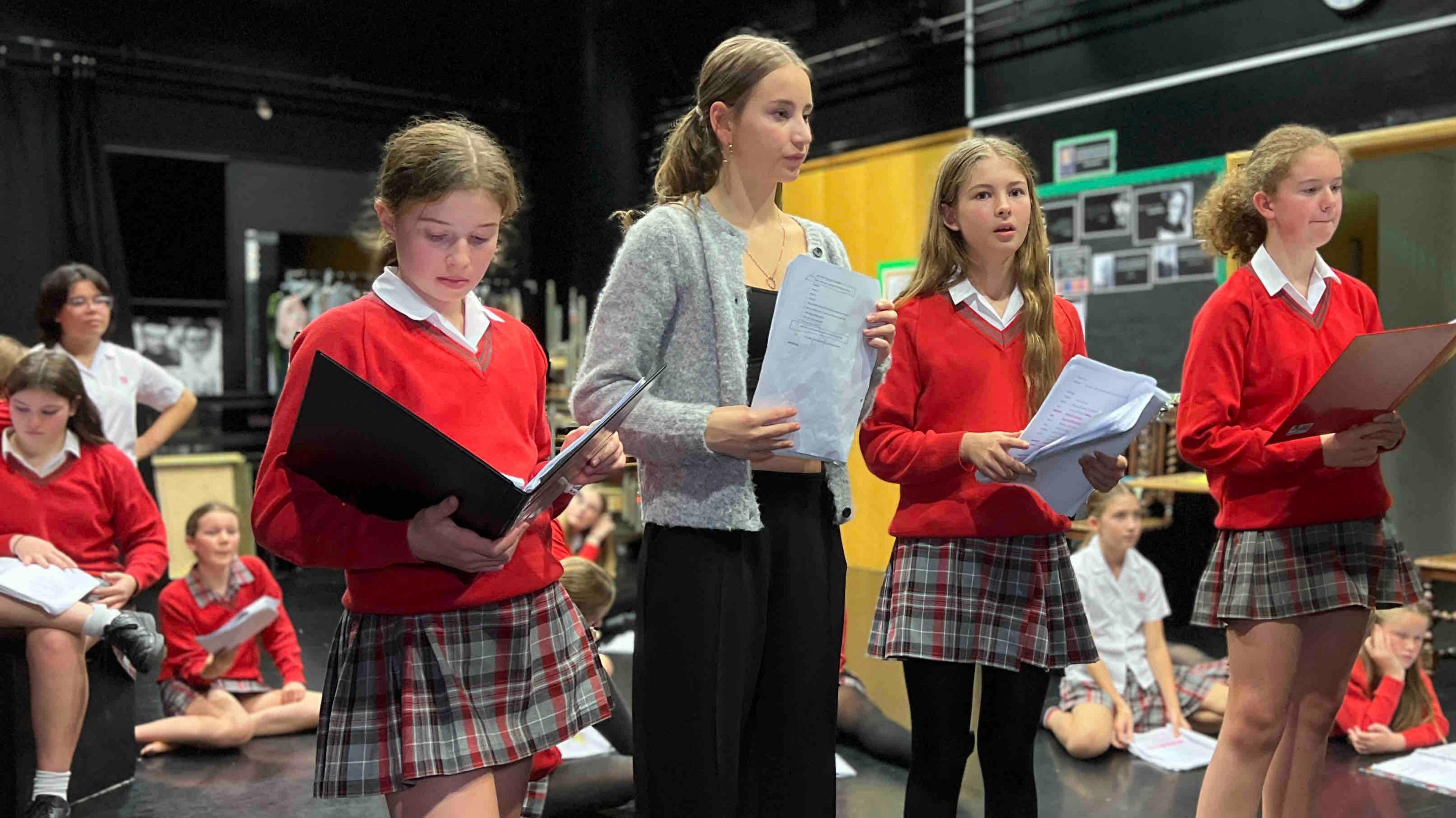 A group of four children standing in a rehearsal space, holding scripts. Three are wearing red sweaters and plaid skirts, while one is wearing black trousers and a grey cardigan. Several others are seated on the floor in the background.