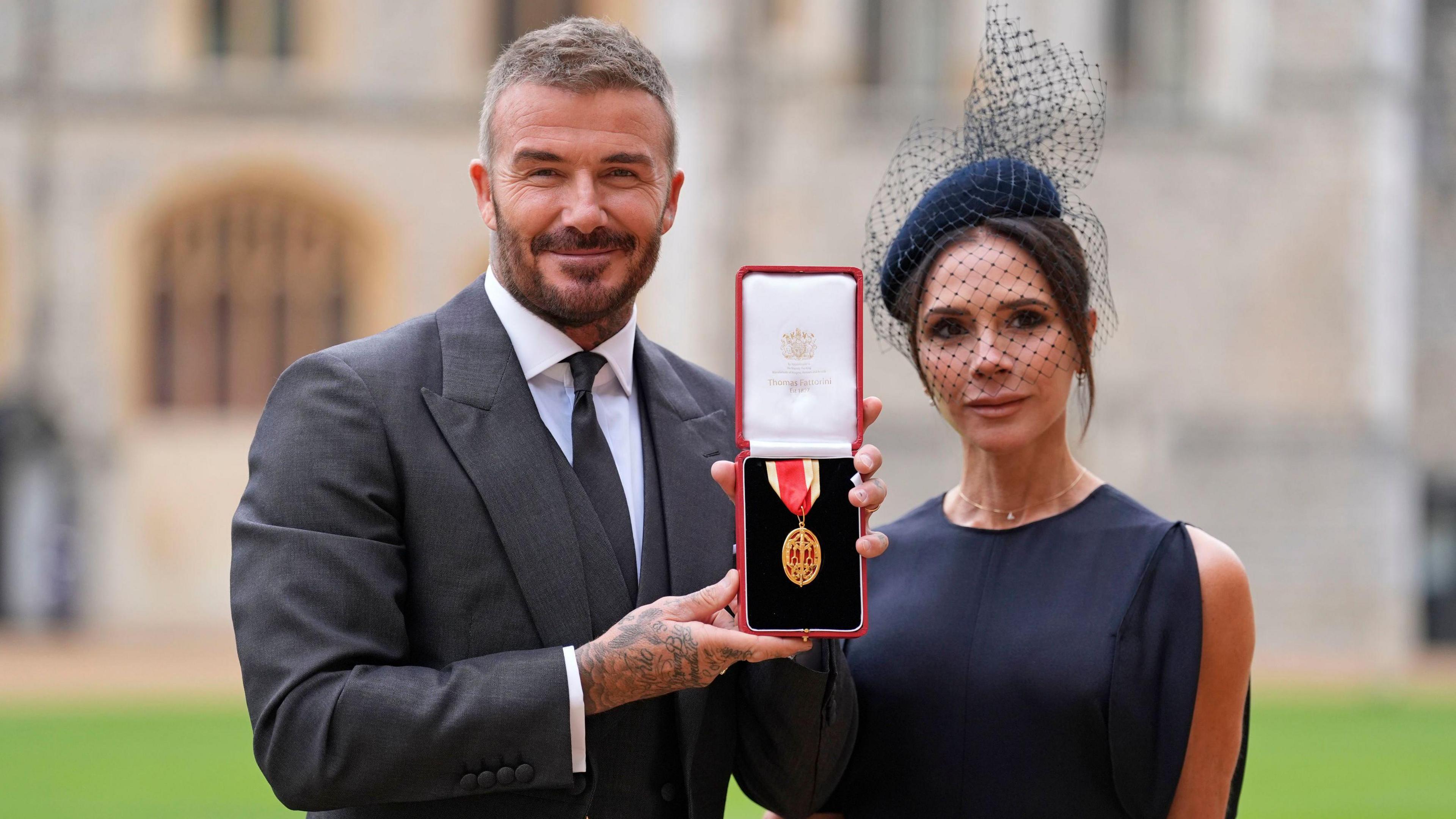 Sir David Beckham with his wife Lady Victoria Beckham, after he was knighted by King Charles at Windsor Castle on Tuesday 4 November. He wears a grey three-piece suit and tie with a white shirt. Victoria Beckham wears a navy blue dress with a matching hat with netting.